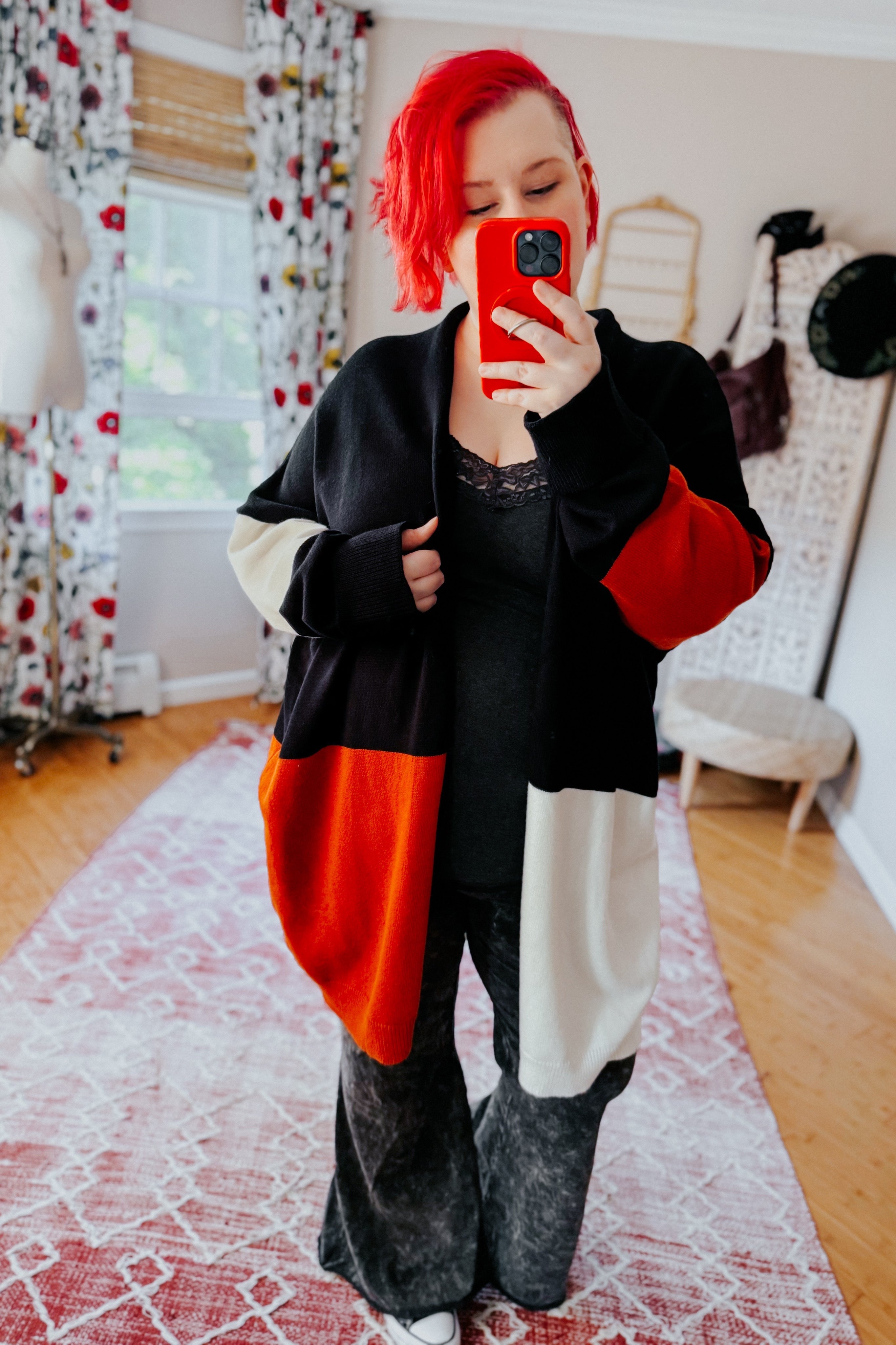Woman with bright red hair wearing a color block open front cardigan in black, red, and cream taking mirror selfie indoors