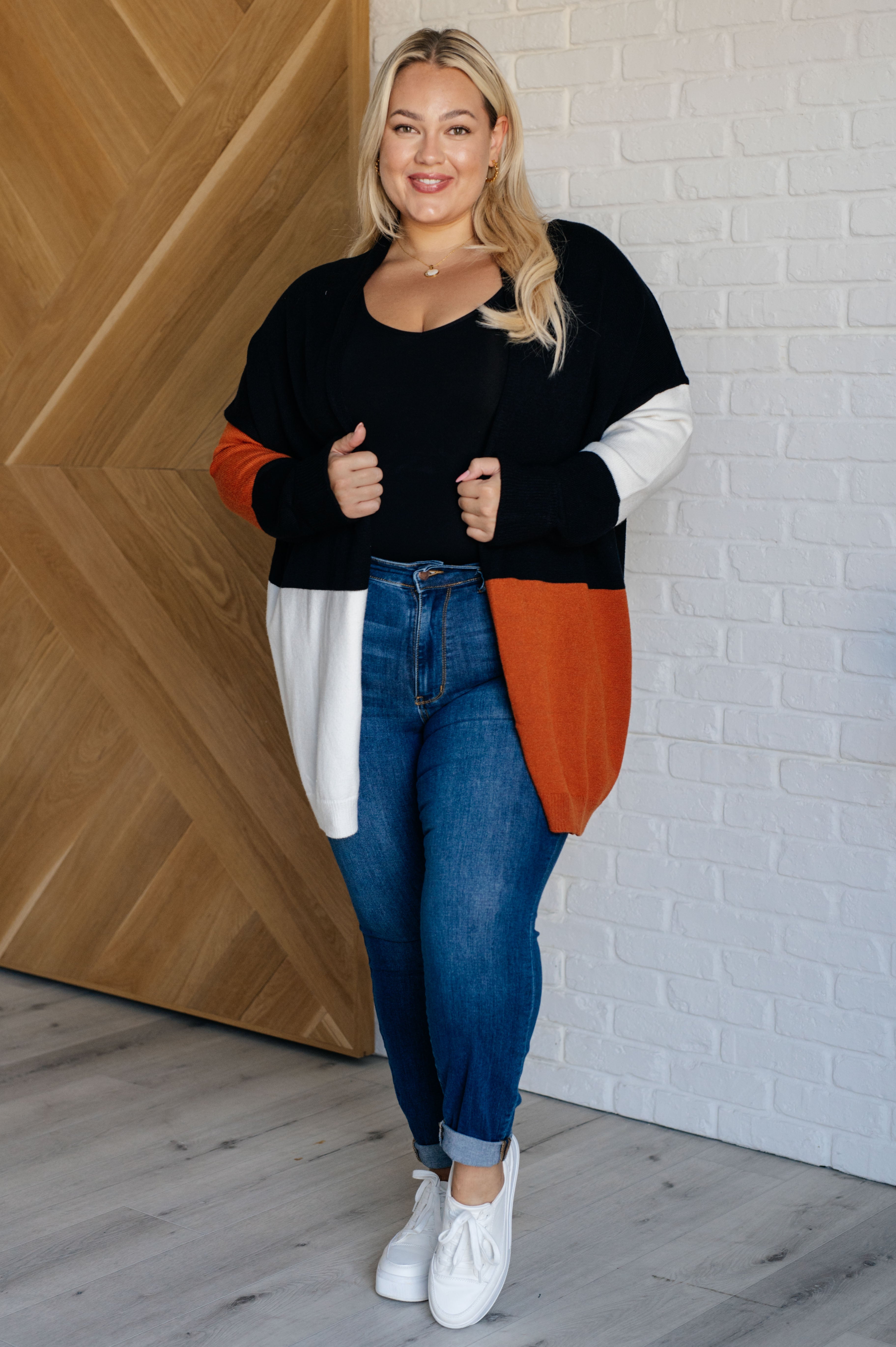Curvy woman wearing color block open front cardigan in black, orange, and white with blue jeans and white sneakers indoors