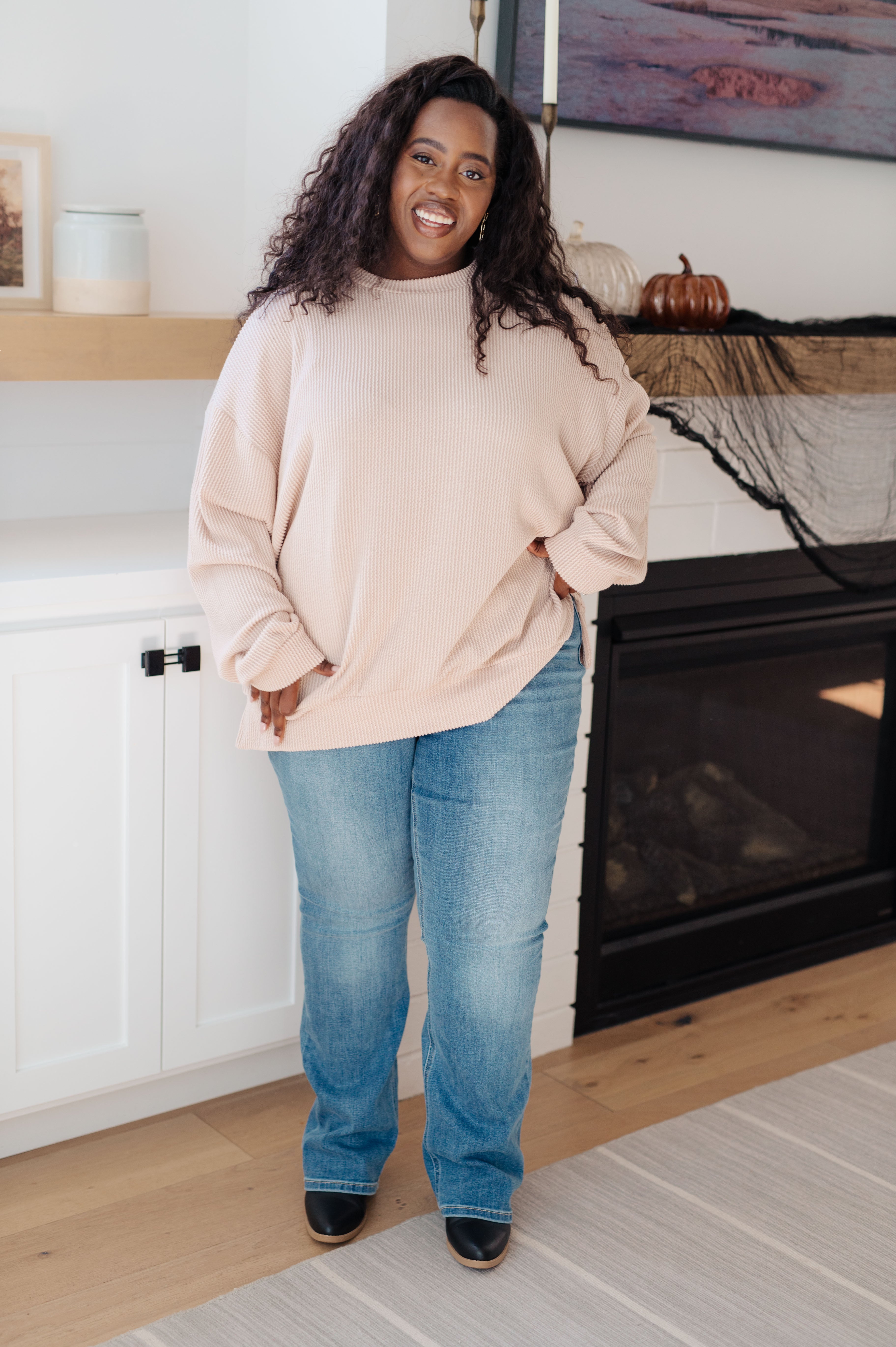 Curvy woman wearing a beige oversized pullover with blue jeans and black boots in cozy modern living room setting