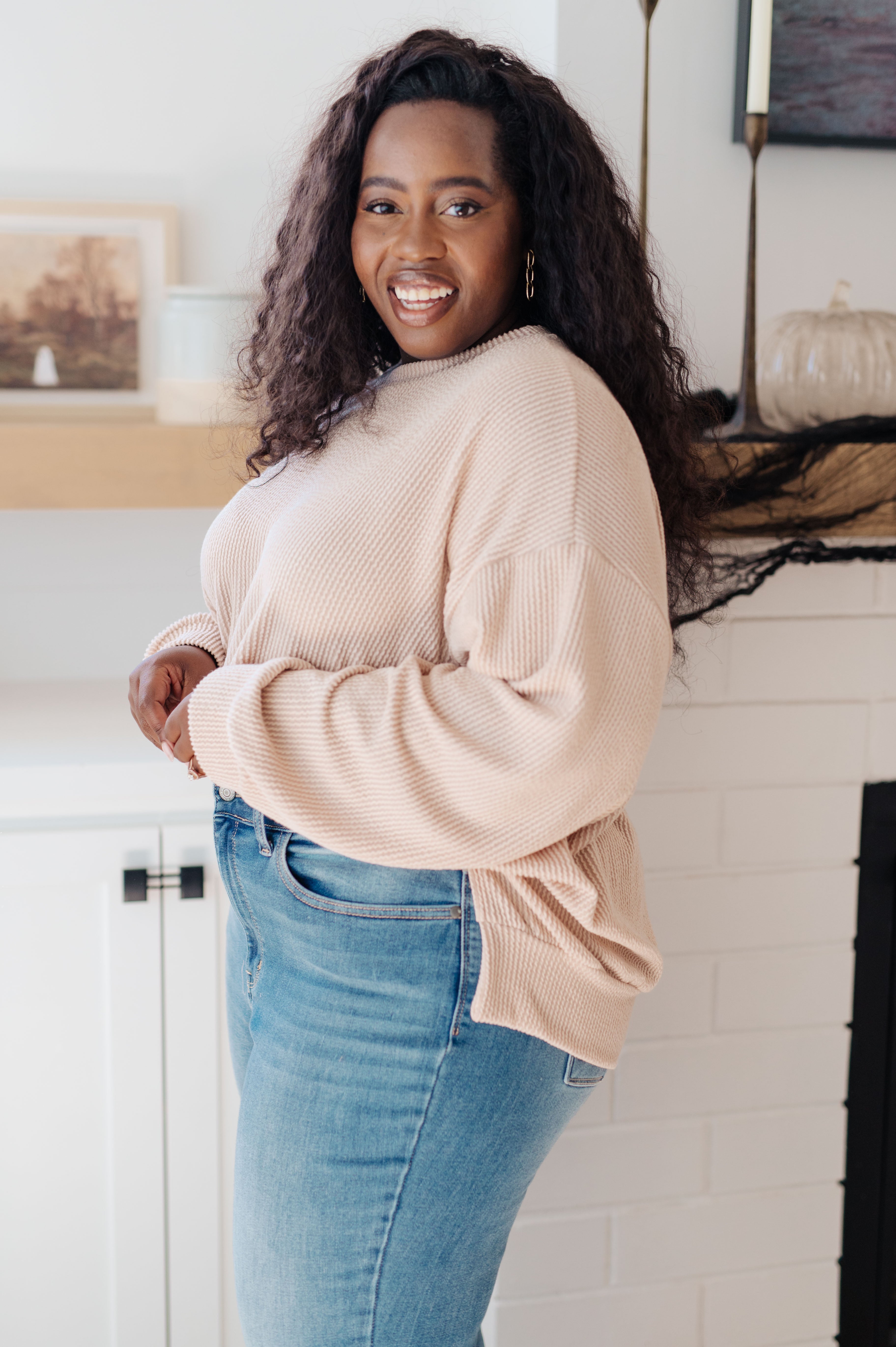 Smiling woman wearing beige oversized pullover sweater paired with light blue jeans in cozy indoor setting