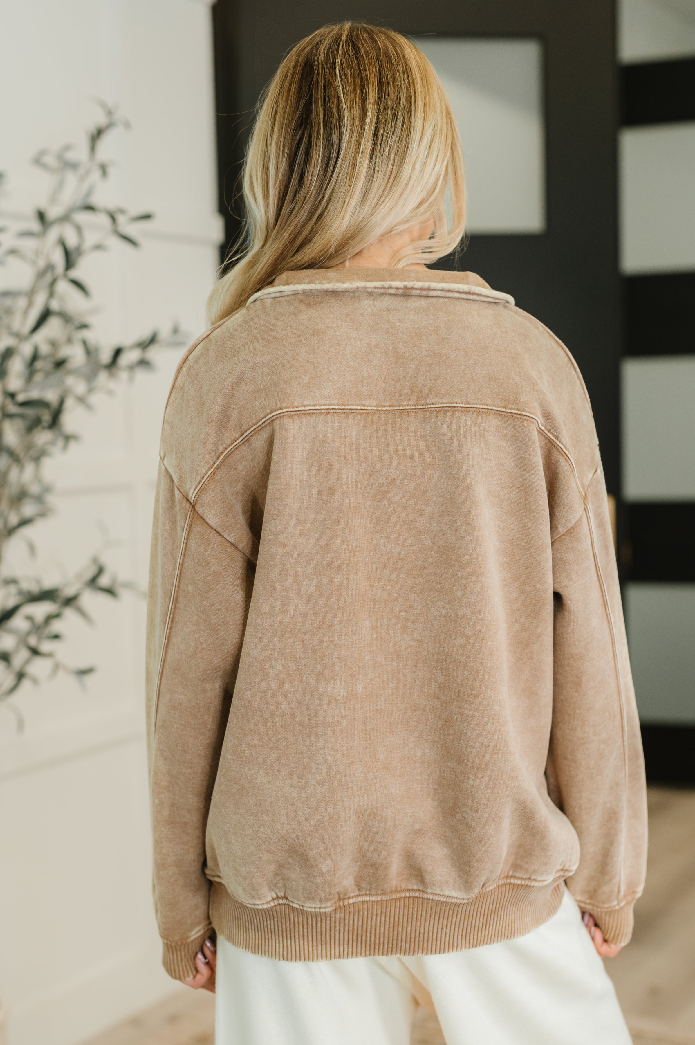 Woman with blonde hair wearing a camel-colored plush half zip pullover viewed from the back indoors