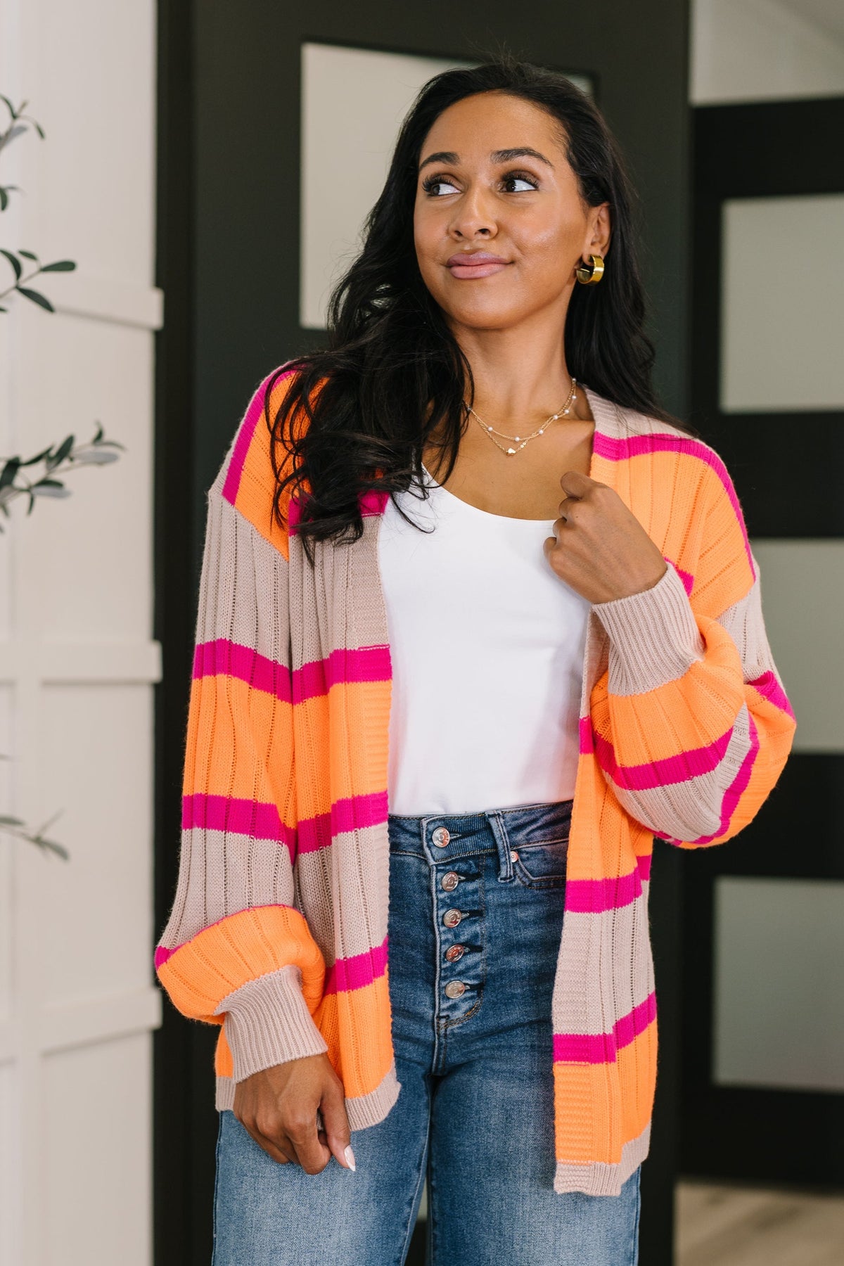 Woman wearing an Orange striped open front cardigan over a white tank top with button fly jeans in a bright modern interior