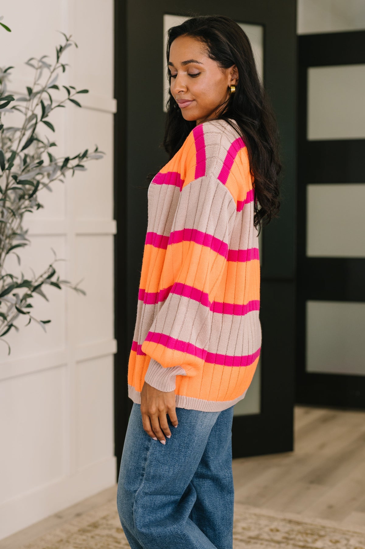Woman modeling an orange striped open front cardigan with dropped shoulders and blue jeans in a modern home entryway