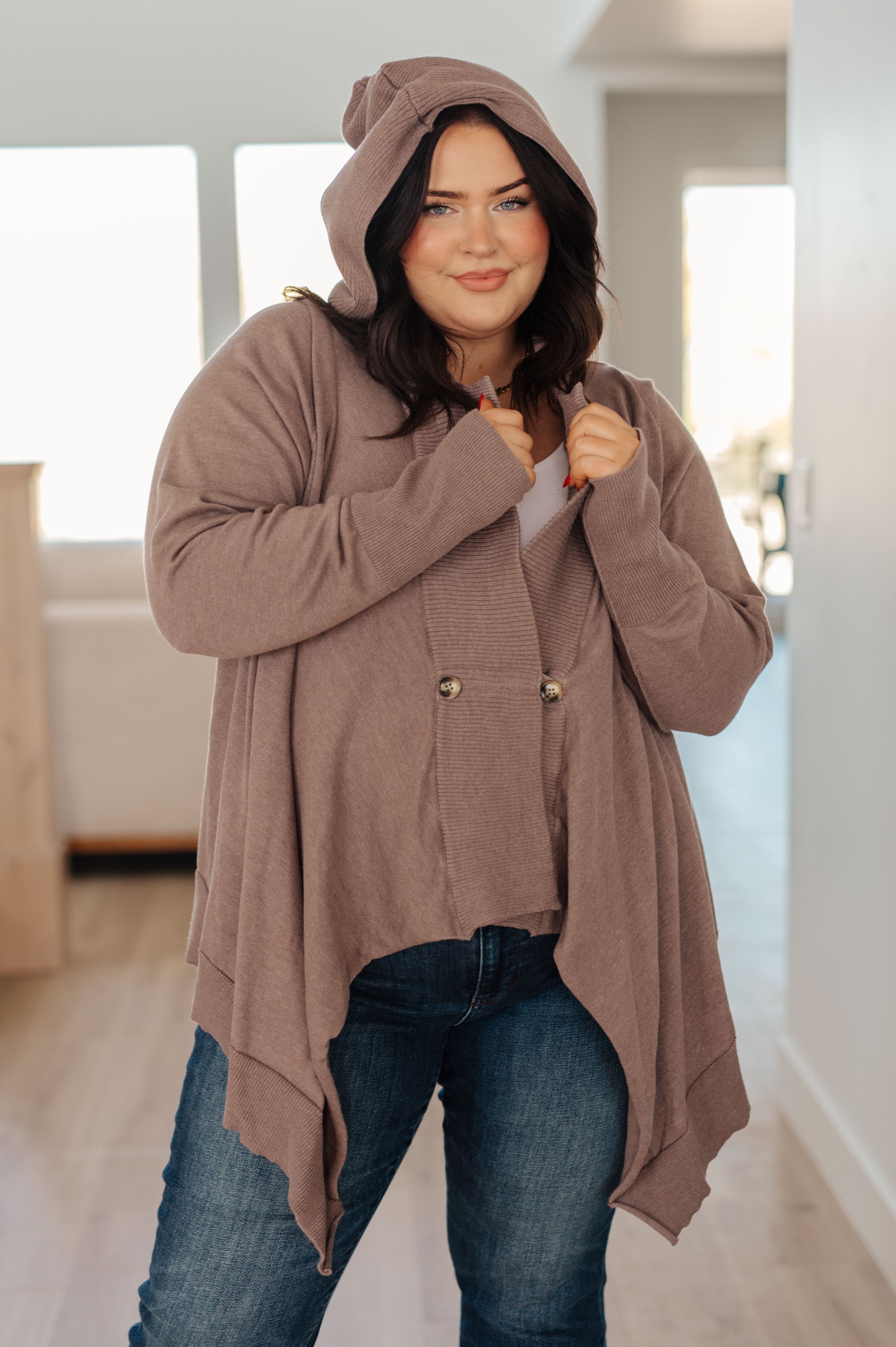 Woman wearing mocha-colored cozy cardigan with hood and asymmetrical hem paired with casual blue jeans indoors
