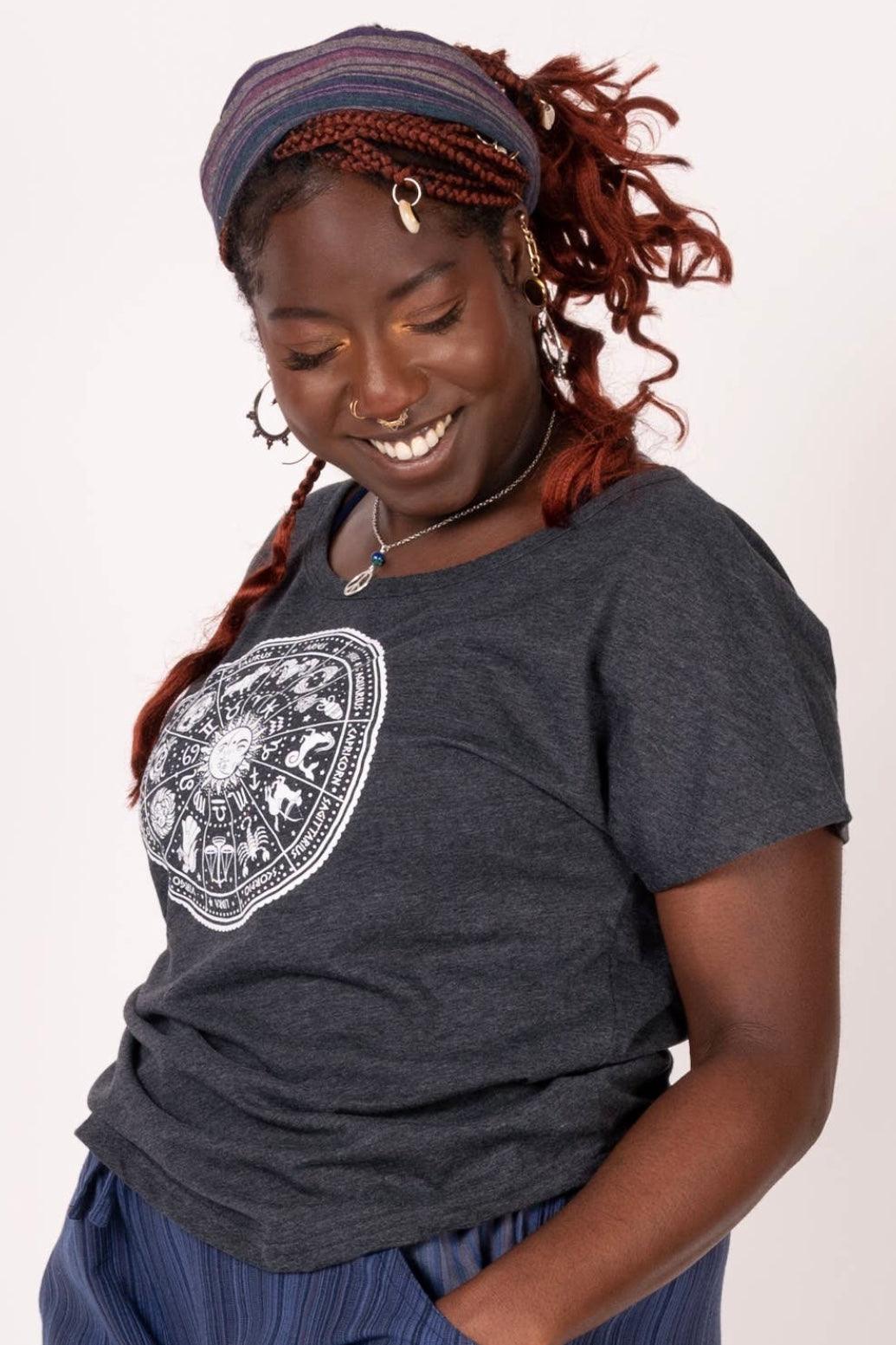 Curly-haired woman wearing charcoal slouchy top with zodiac graphic smiling and looking down in studio setting