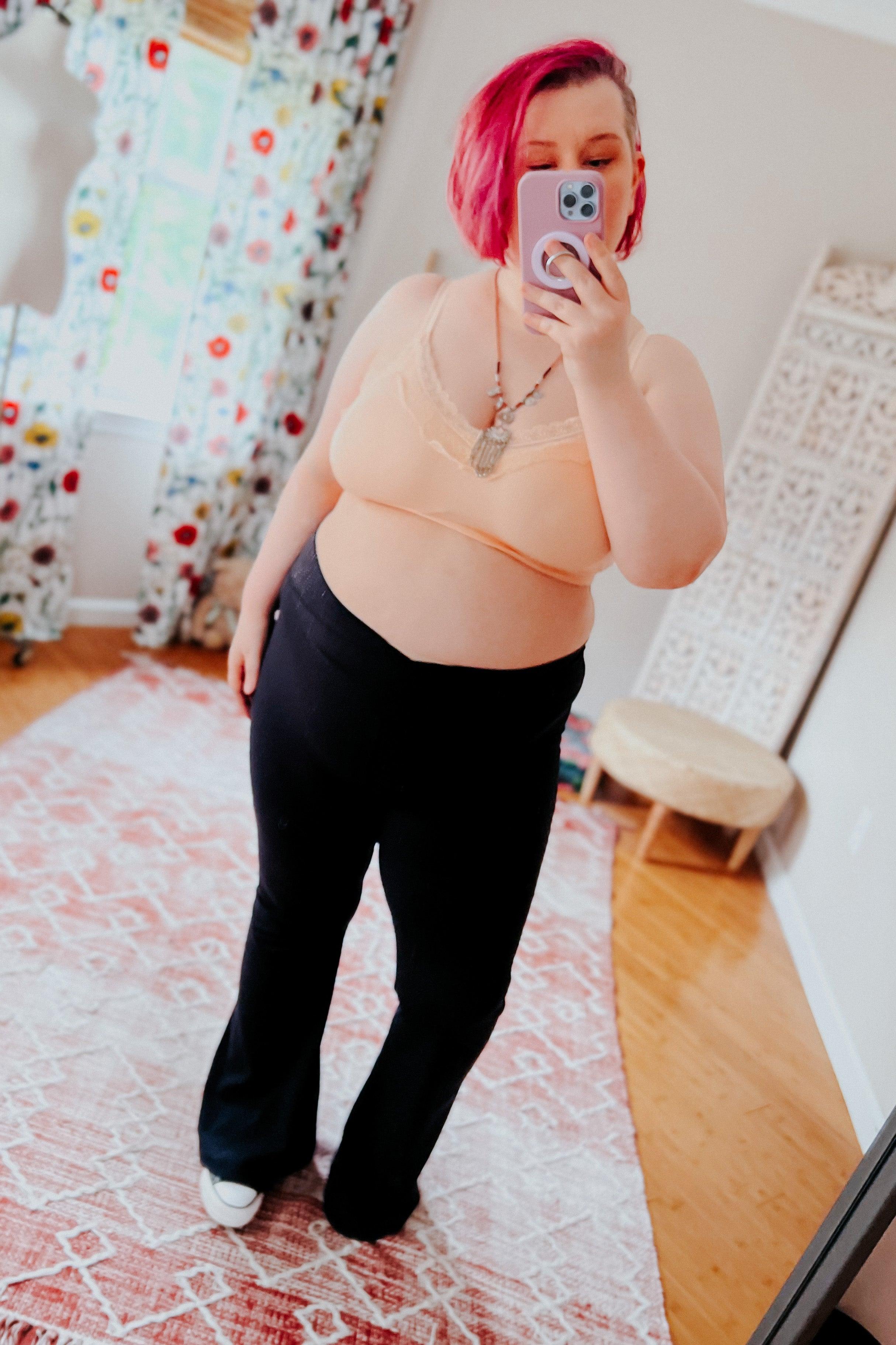 Plus-size woman with short pink hair wearing peach lace-trim bralette and black pants taking mirror selfie in bright room