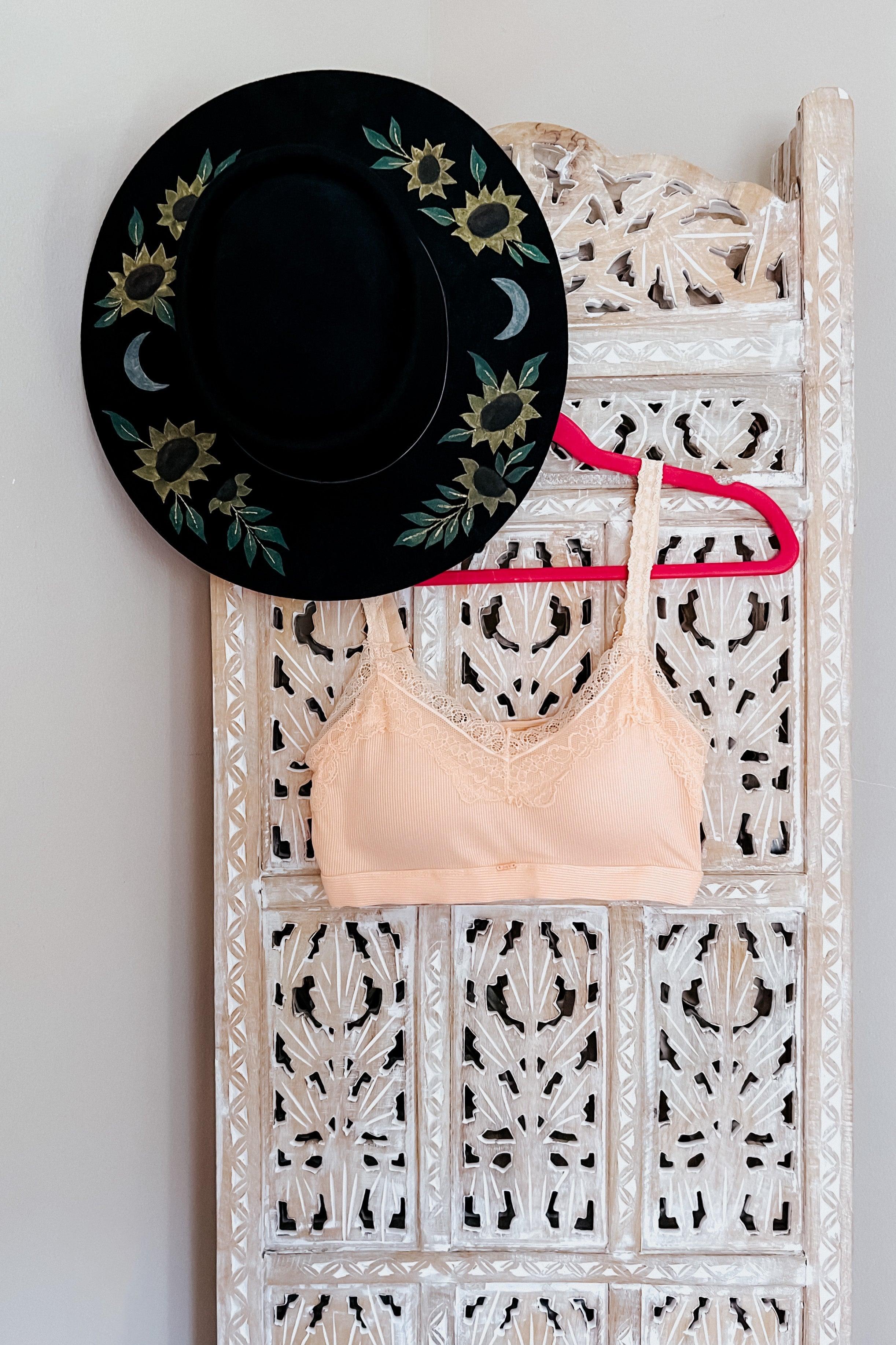 Peach lace-trim bralette hanging on carved wooden screen with black hat decorated with sunflowers and moons
