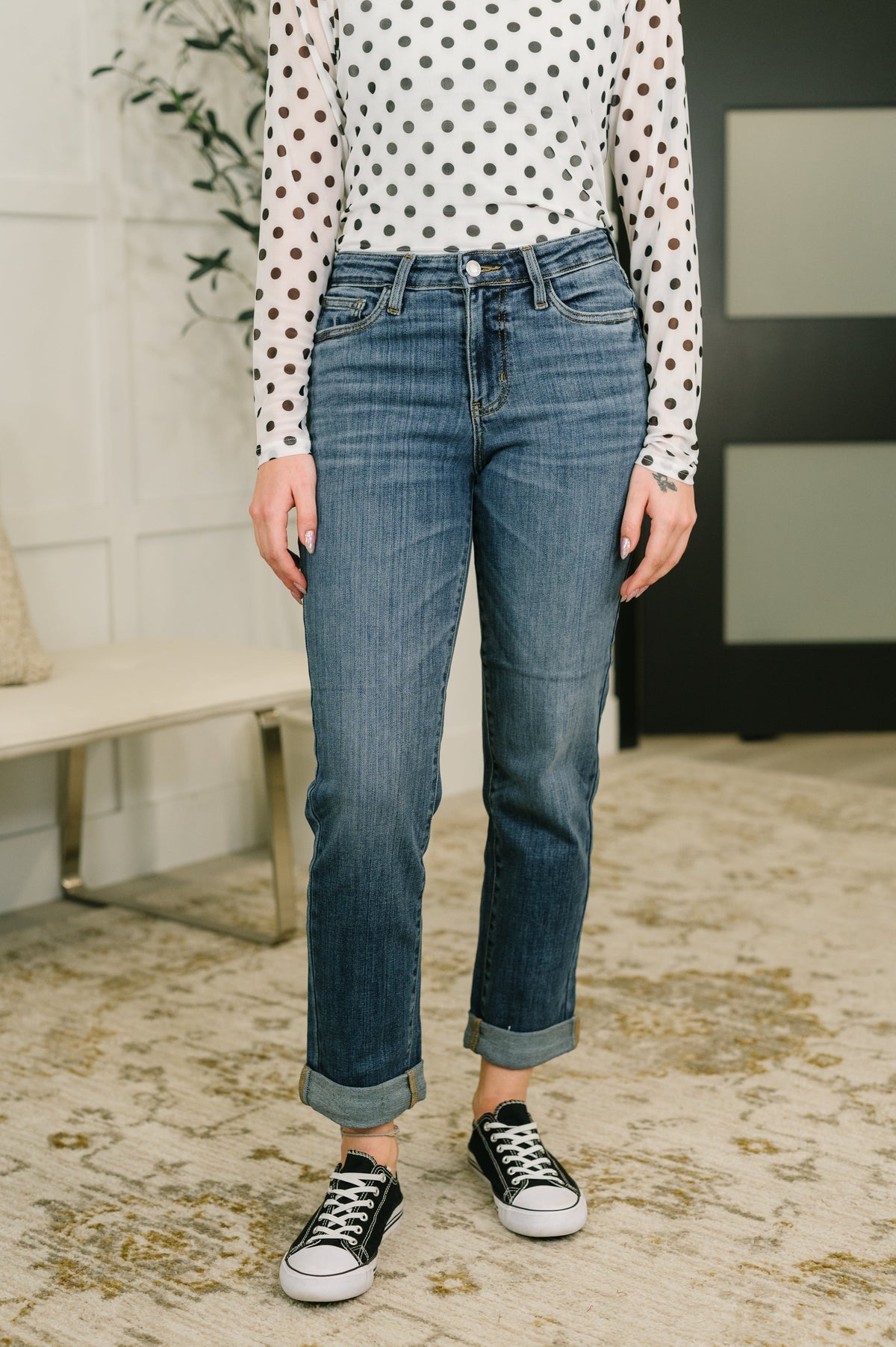 Woman wearing blue mid-rise boyfriend jeans with cuffed hems and a relaxed fit, paired with a polka dot top and sneakers indoors