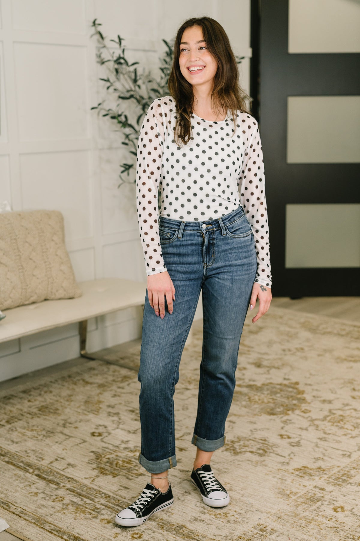 Woman wearing blue mid-rise boyfriend jeans with a white polka dot top and black sneakers indoors