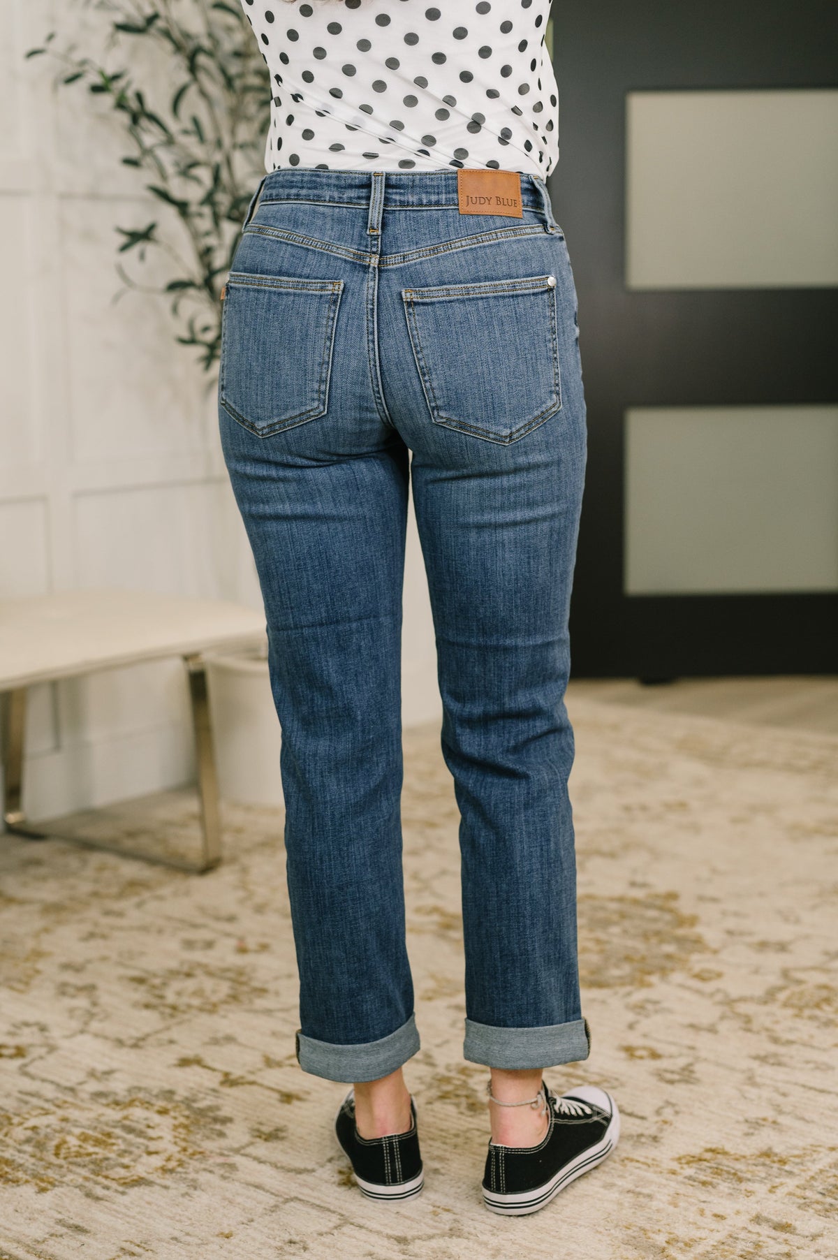 Back view of a woman wearing blue mid rise boyfriend jeans with cuffed hems and a polka dot top styled with black sneakers indoors