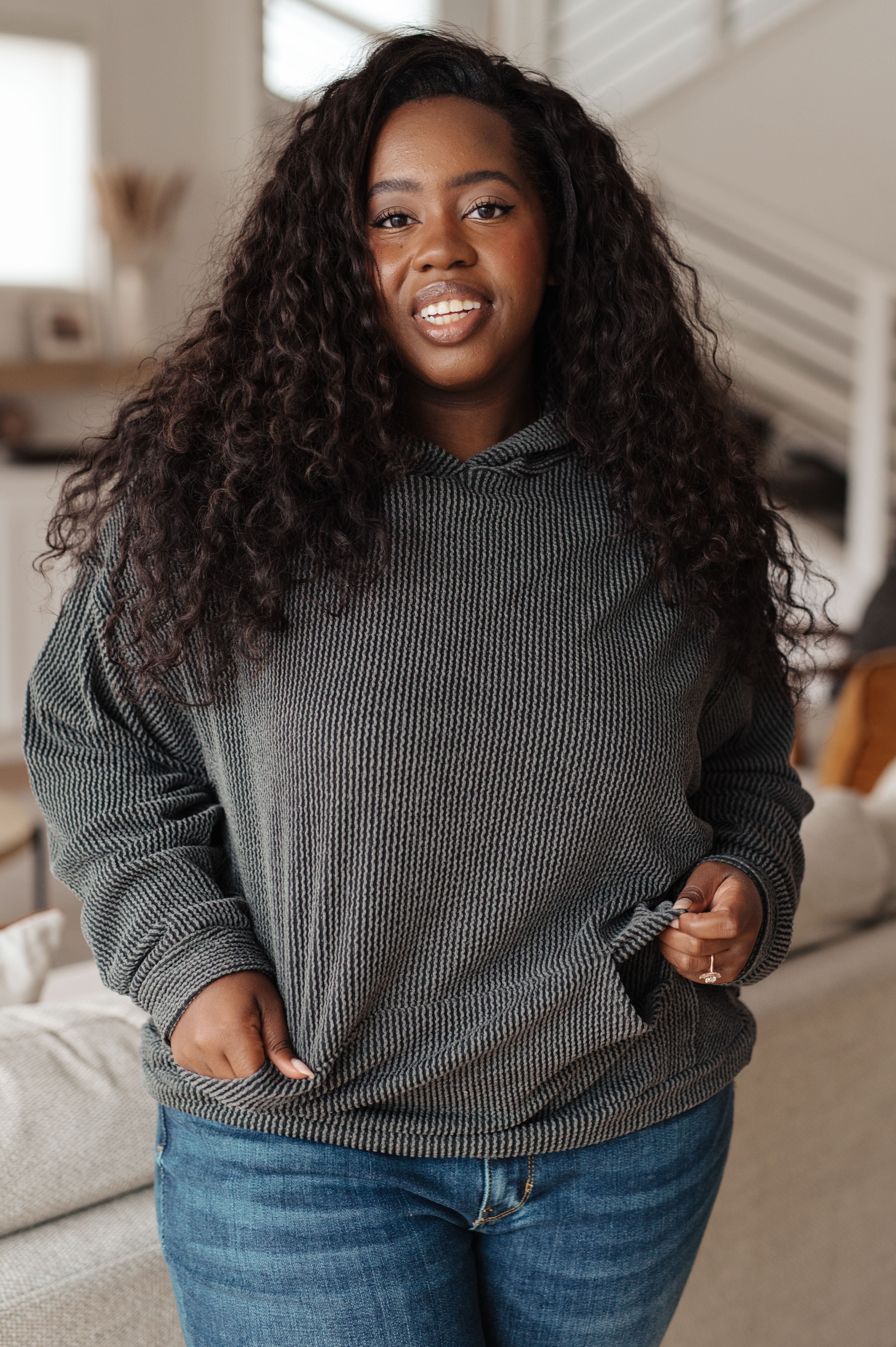 Woman wearing a grey rib knit hoodie over blue jeans against an indoor setting.