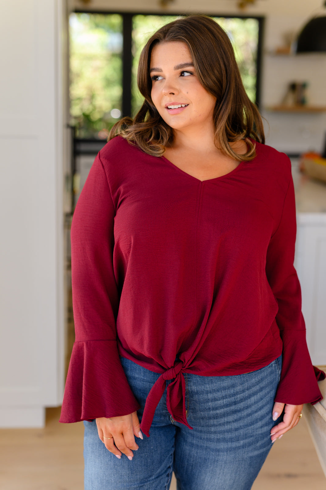 Curvy woman indoors wearing deep red long sleeve top with flare cuffs and front tie paired with blue jeans