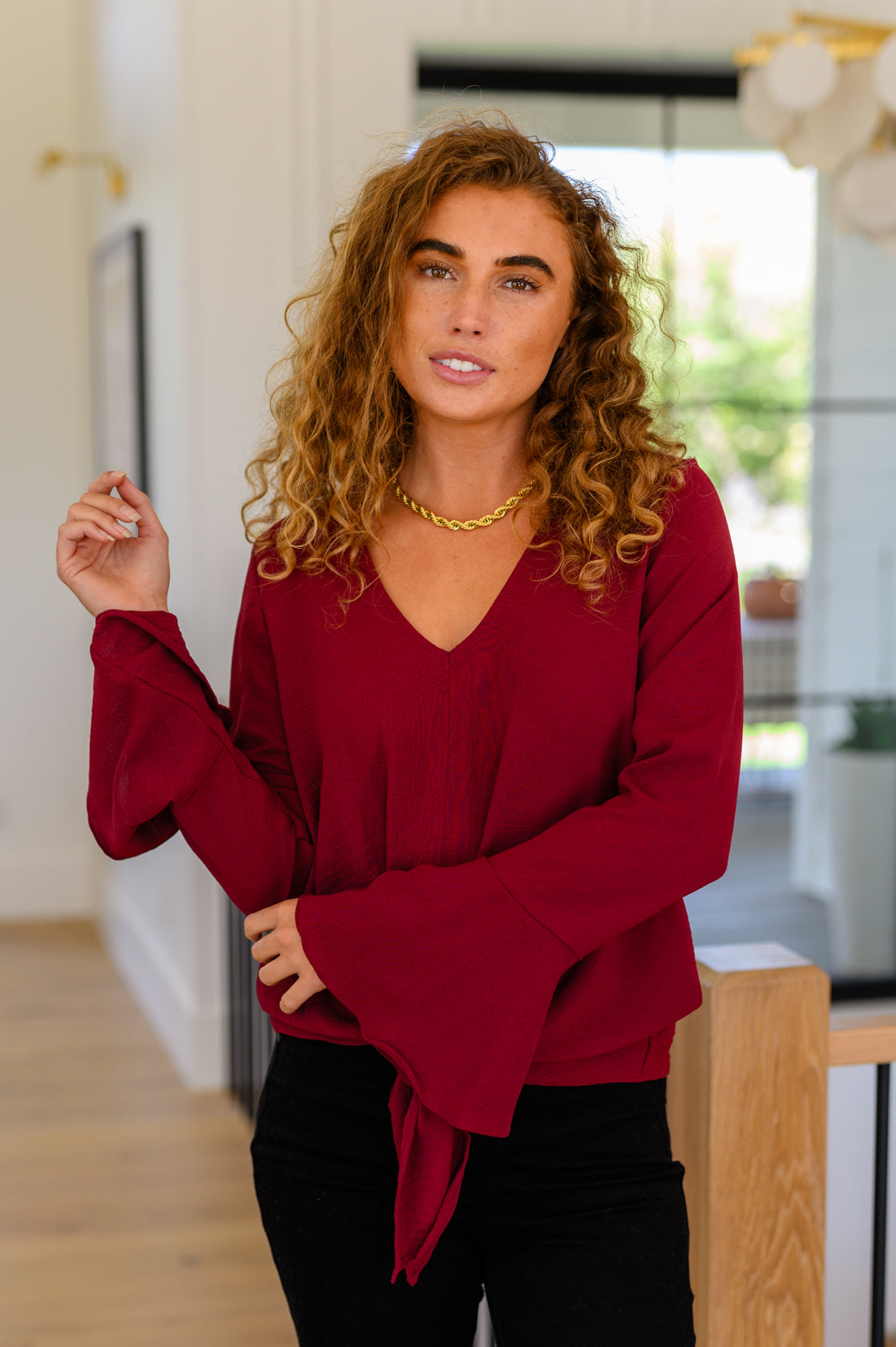 Woman with curly hair wearing deep red V-neck top with dramatic flare sleeves and front bow detail indoors