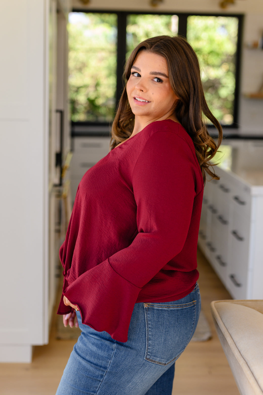 Curvy woman in bright kitchen wearing deep red long sleeve top with flare sleeves and bow detail at hem