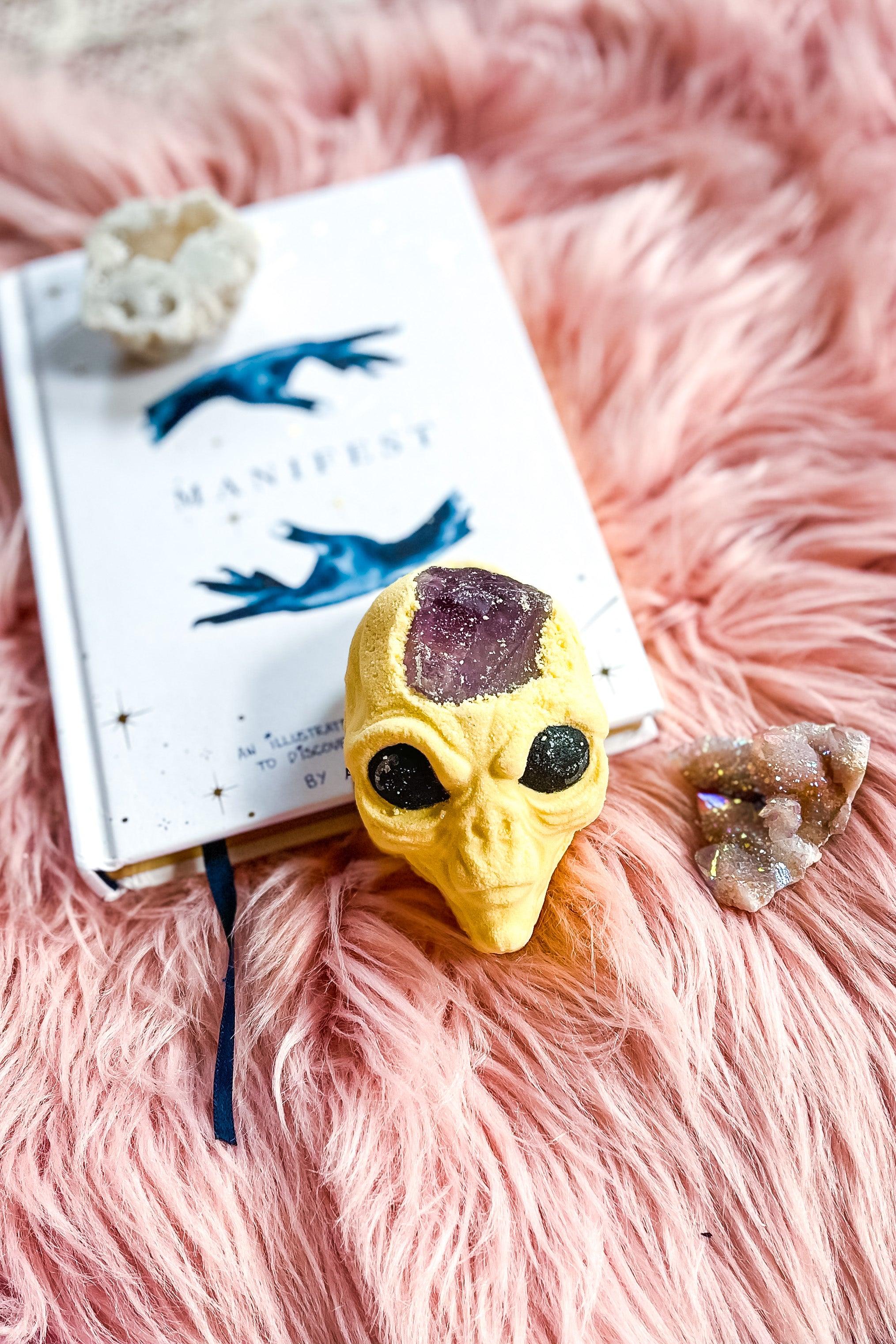 Yellow alien-shaped bath bomb with a purple amethyst crystal top placed on a pink fuzzy surface next to a book and crystals