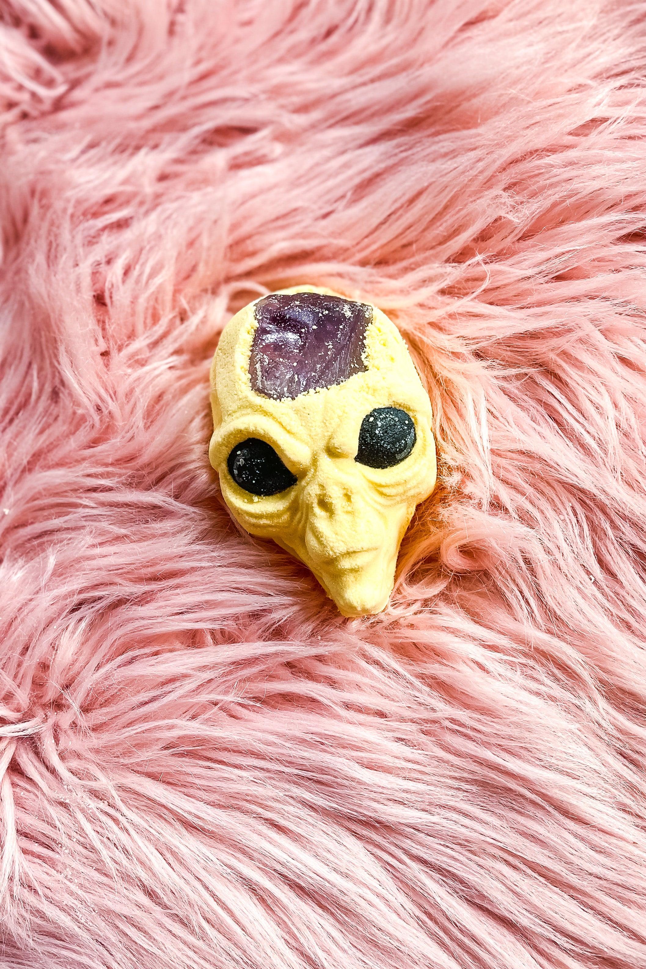Alien-shaped yellow bath bomb with embedded purple amethyst crystal resting on soft pink faux fur background