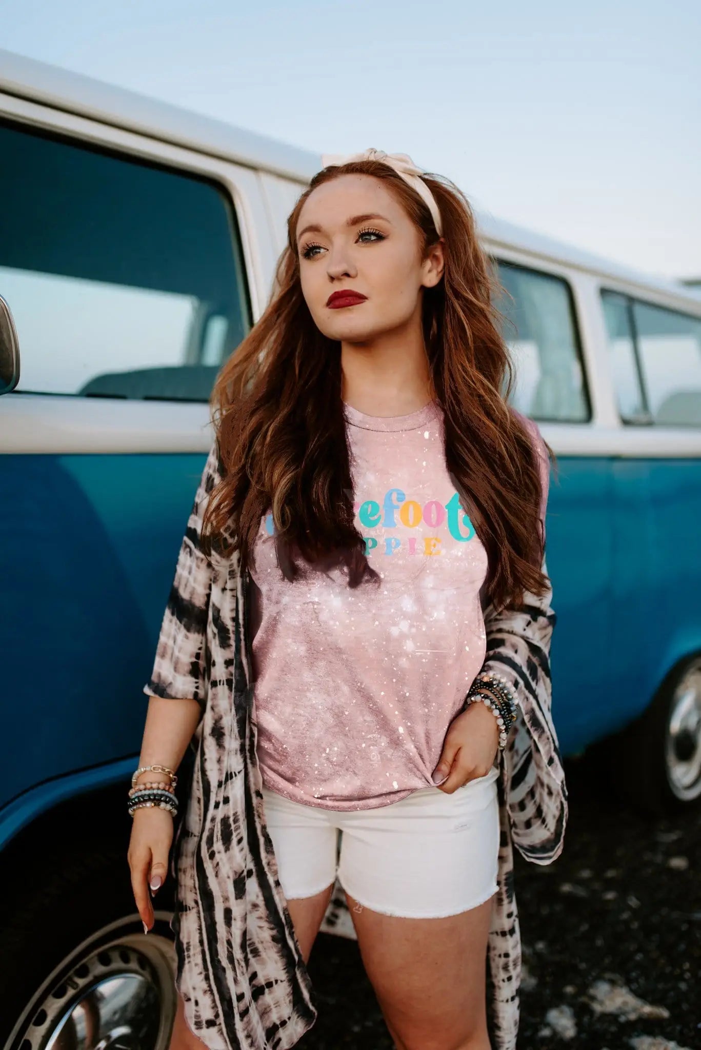Woman wearing barefoot hippie pink bleached tee with colorful text, white shorts, and tie-dye outerwear by blue vintage van