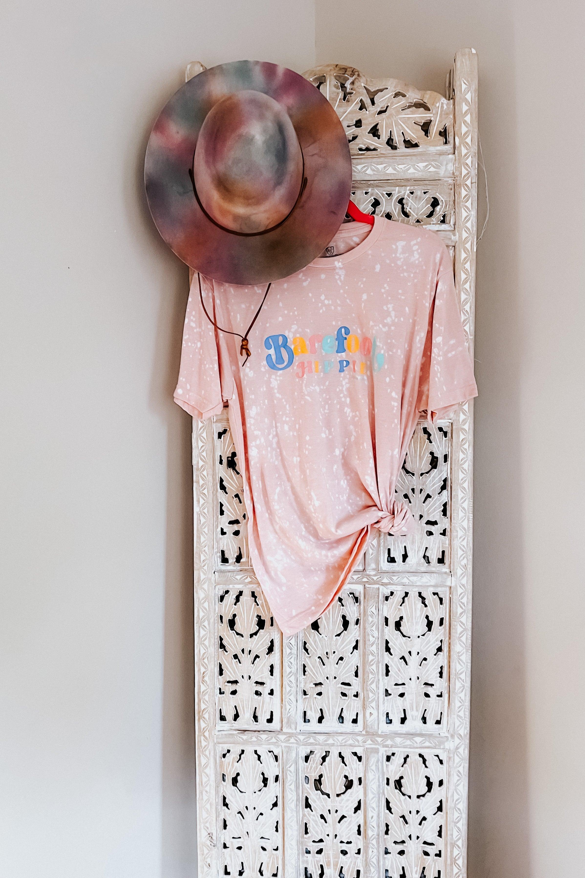 Pink bleached Barefoot Hippie tee with colorful logo knotted on white carved wooden screen with tie-dye hat hanging above