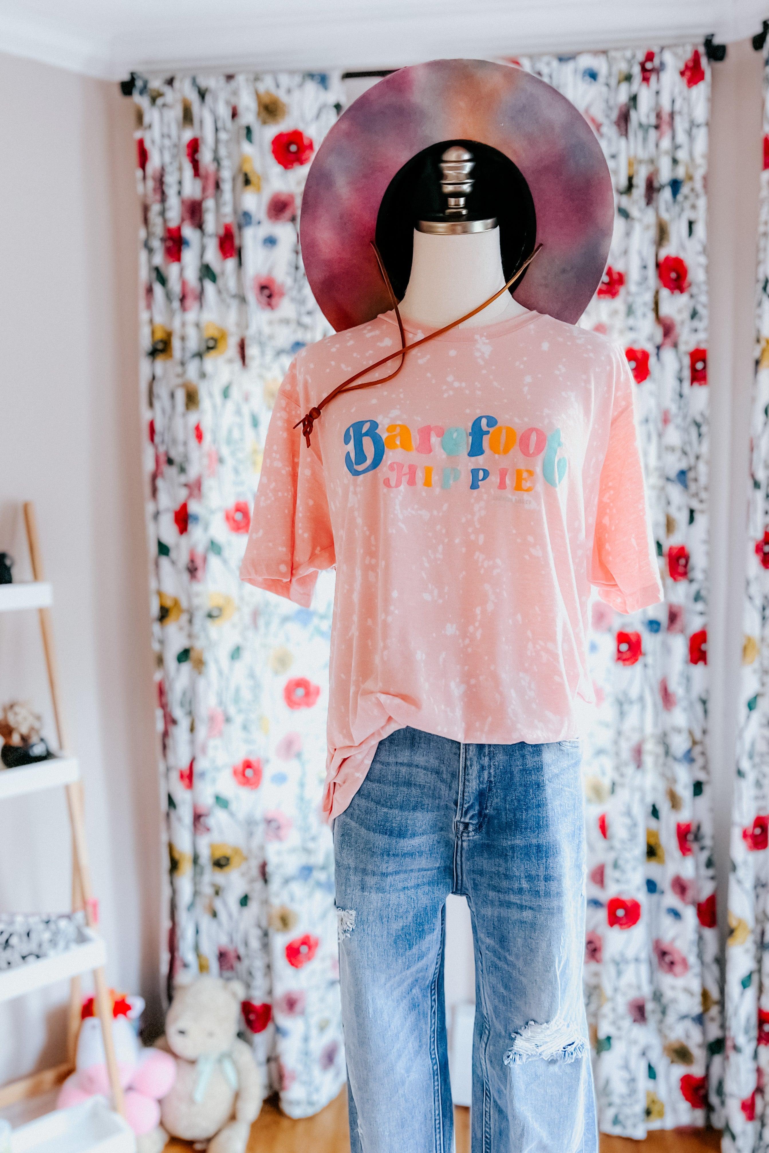Mannequin dressed in barefoot hippie pink bleached tee with colorful text and wide-brim hat indoors by floral curtains