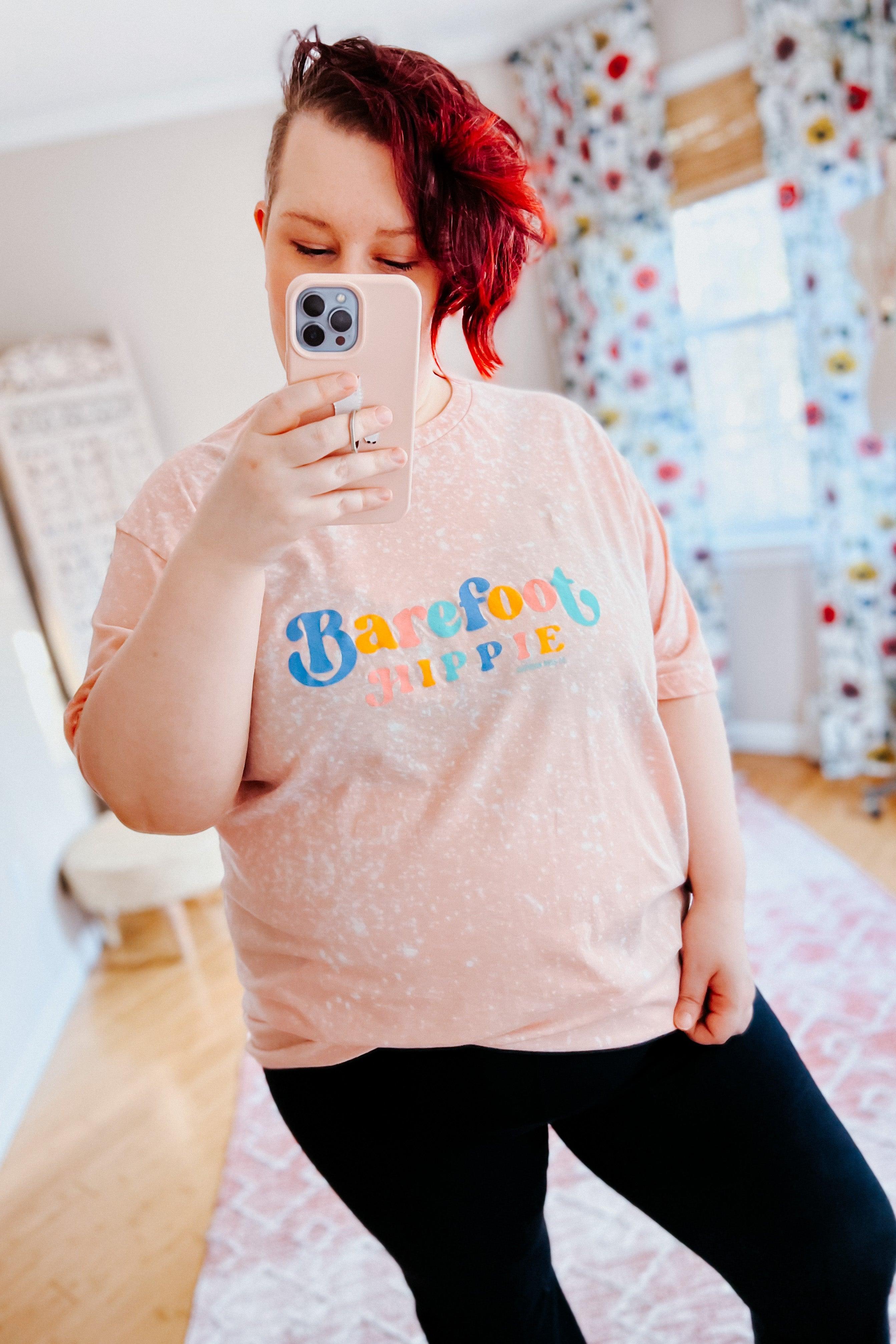 Person with short red hair wearing barefoot hippie pink bleached tee with colorful text taking indoor selfie in bright room