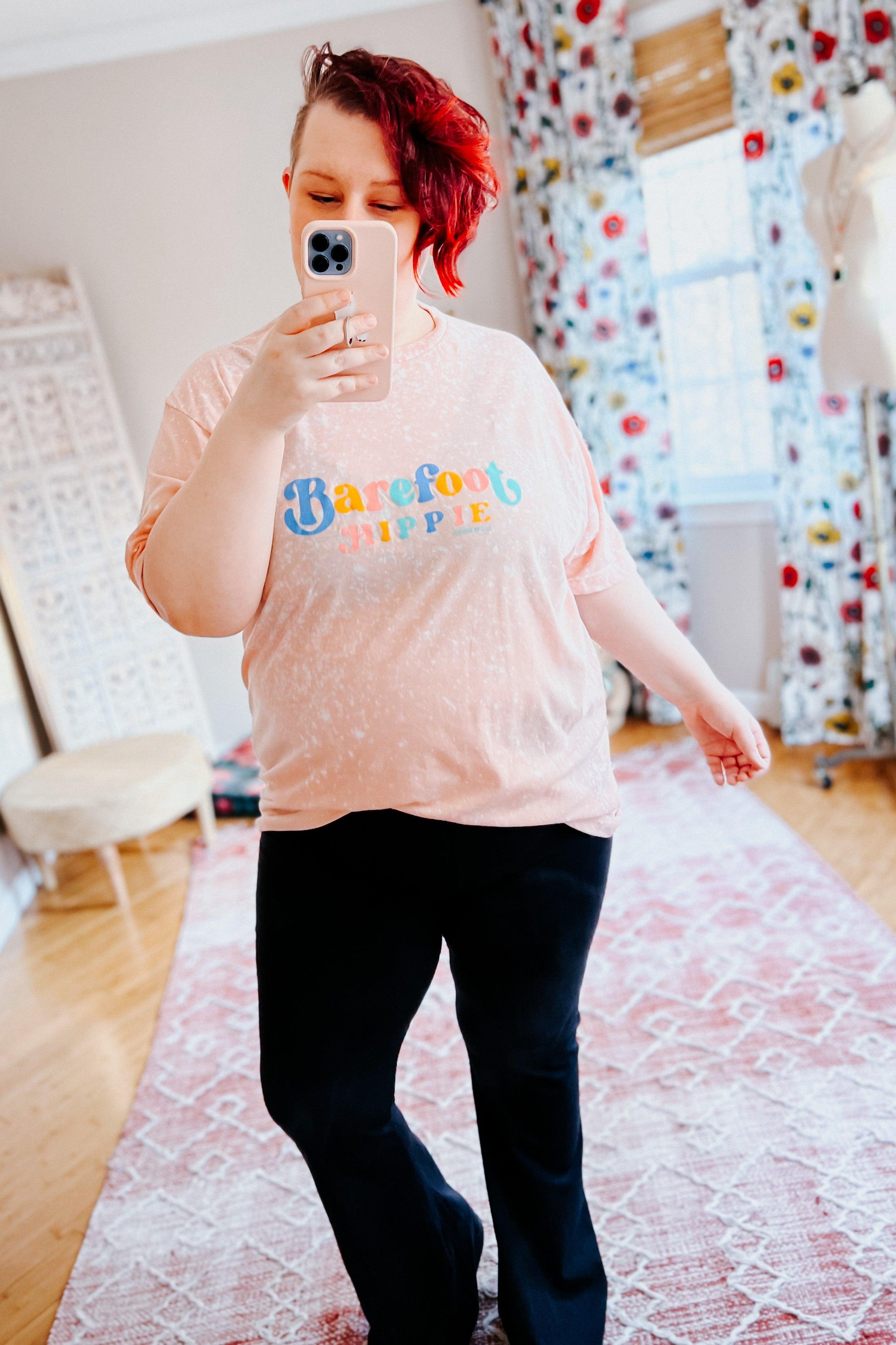 Person with red hair wearing Barefoot Hippie pink bleached tee with colorful text taking mirror selfie in bright room