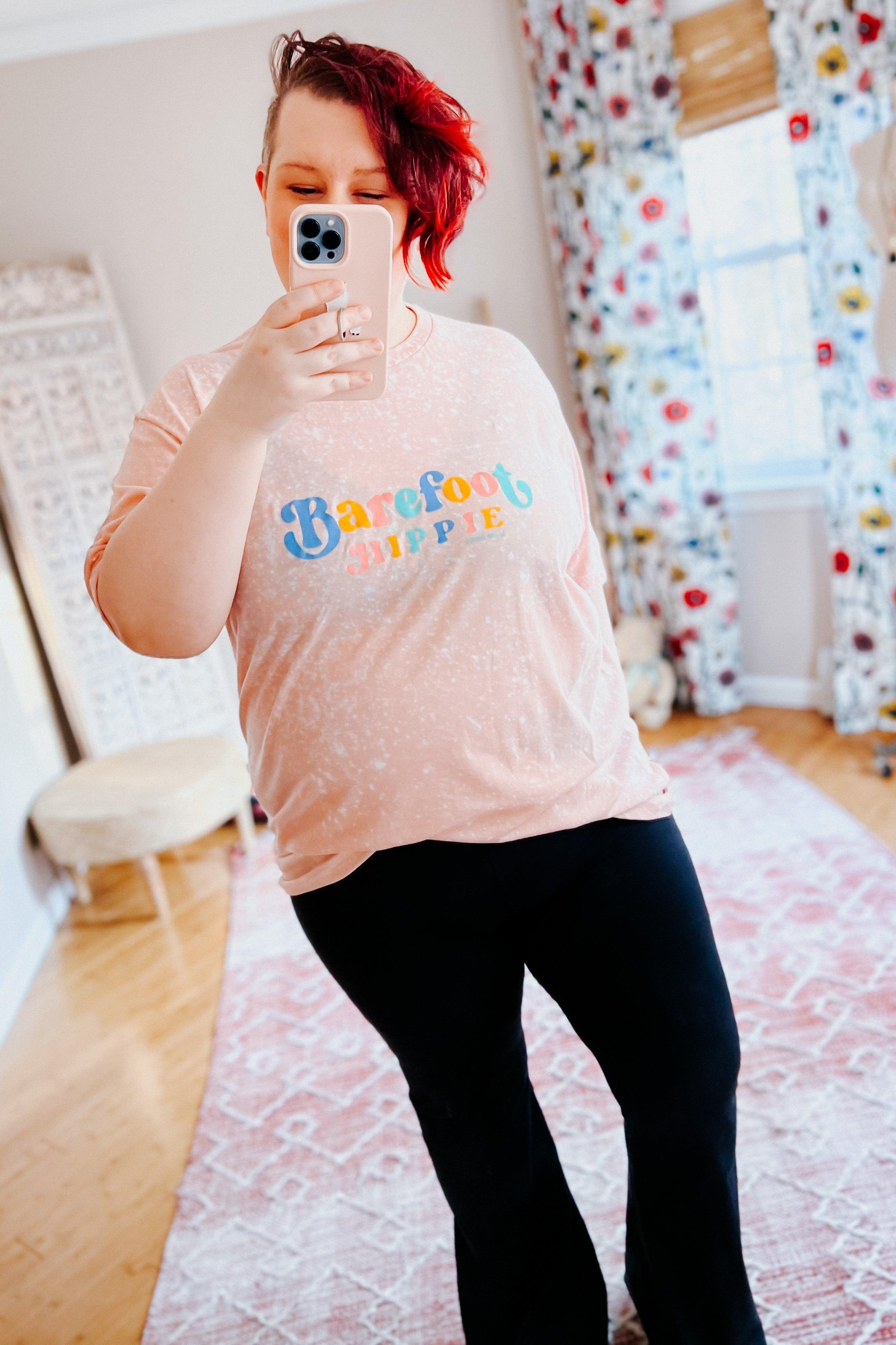 Person with short red hair wearing barefoot hippie pink bleached tee with colorful text taking indoor mirror selfie in bright room