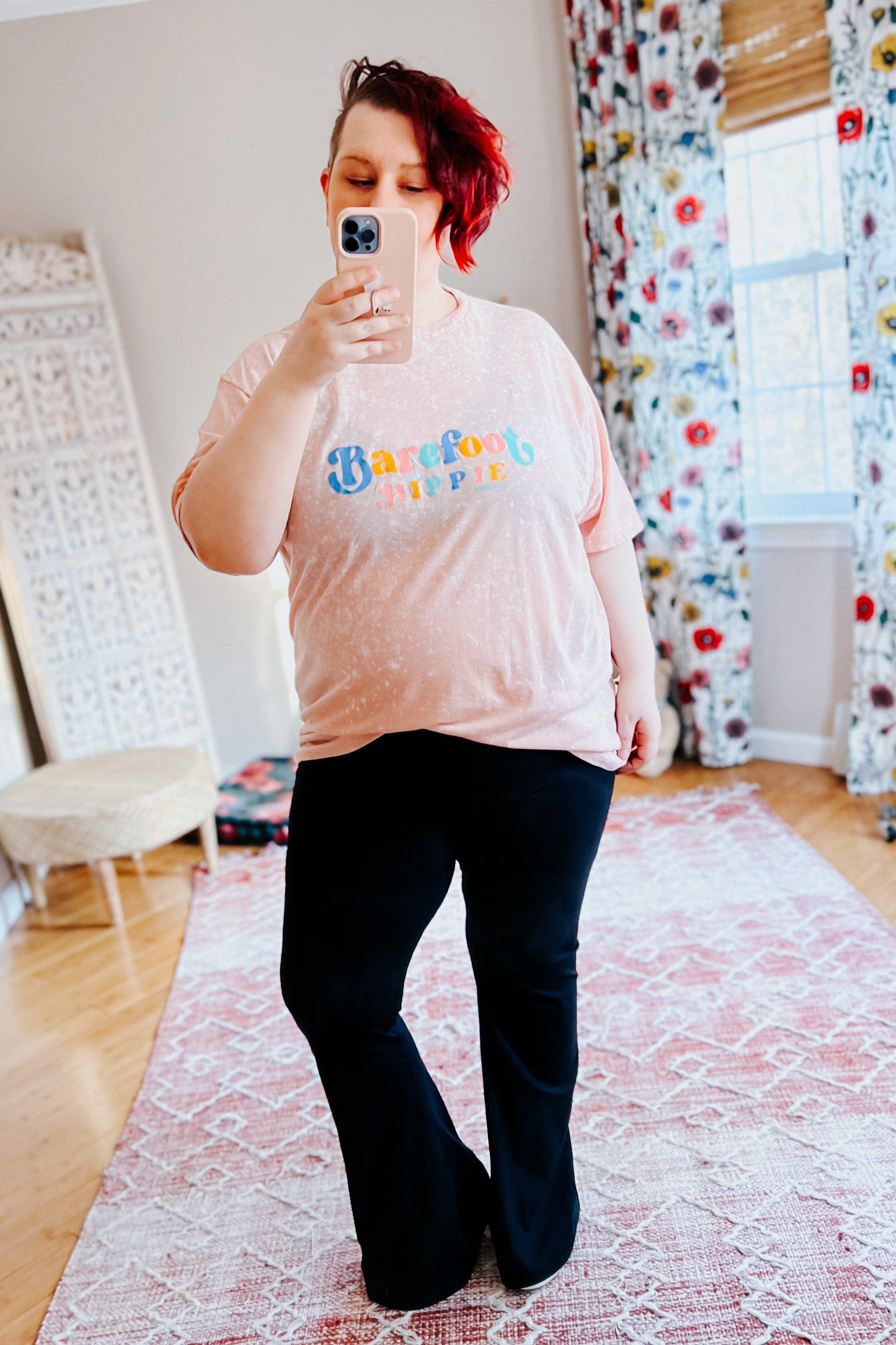 Person with short red hair wearing barefoot hippie pink bleached tee with colorful text taking mirror selfie indoors