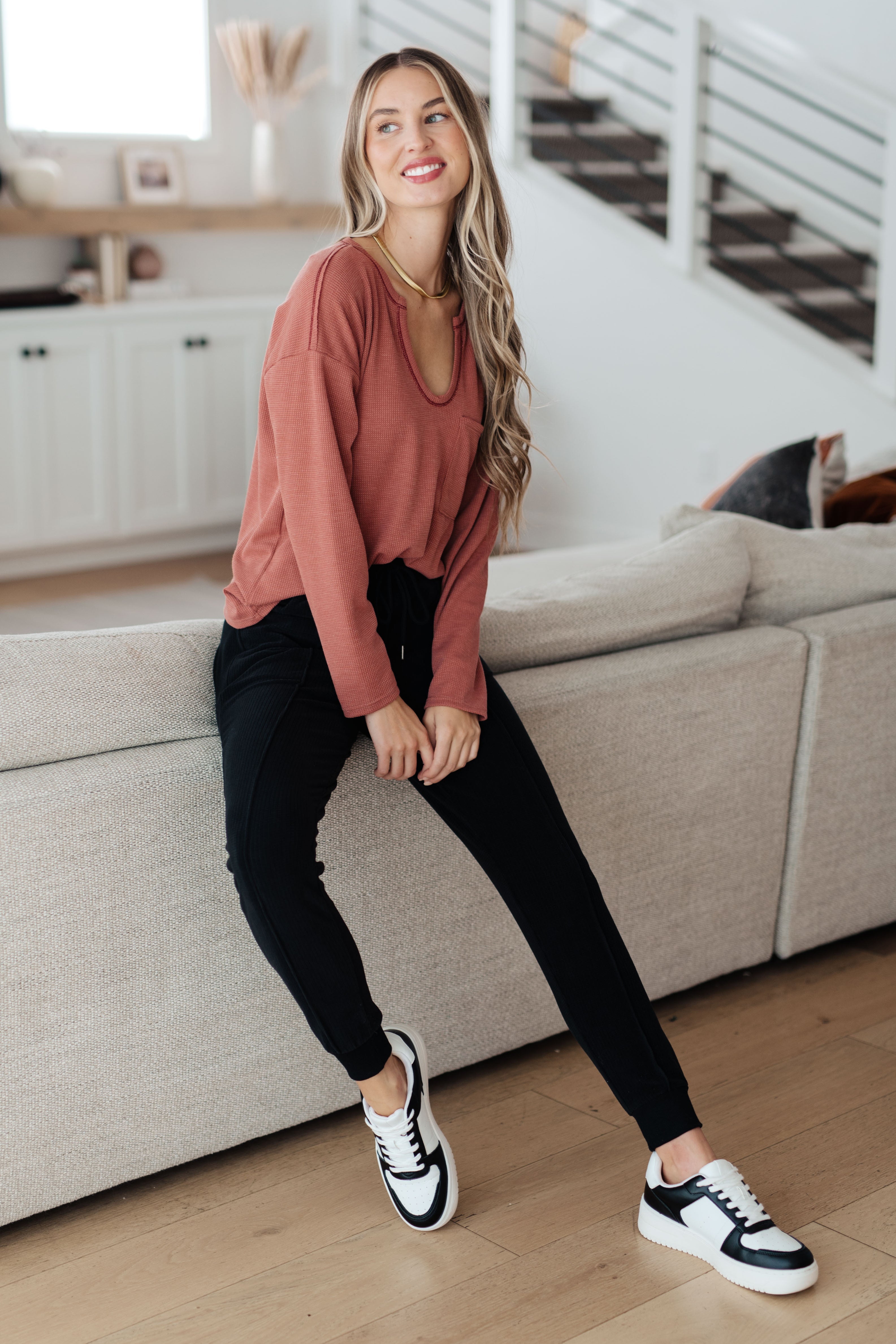 Woman wearing rust-colored Behind The Scenes waffle knit top with scoop neck sitting on a light gray sofa indoors