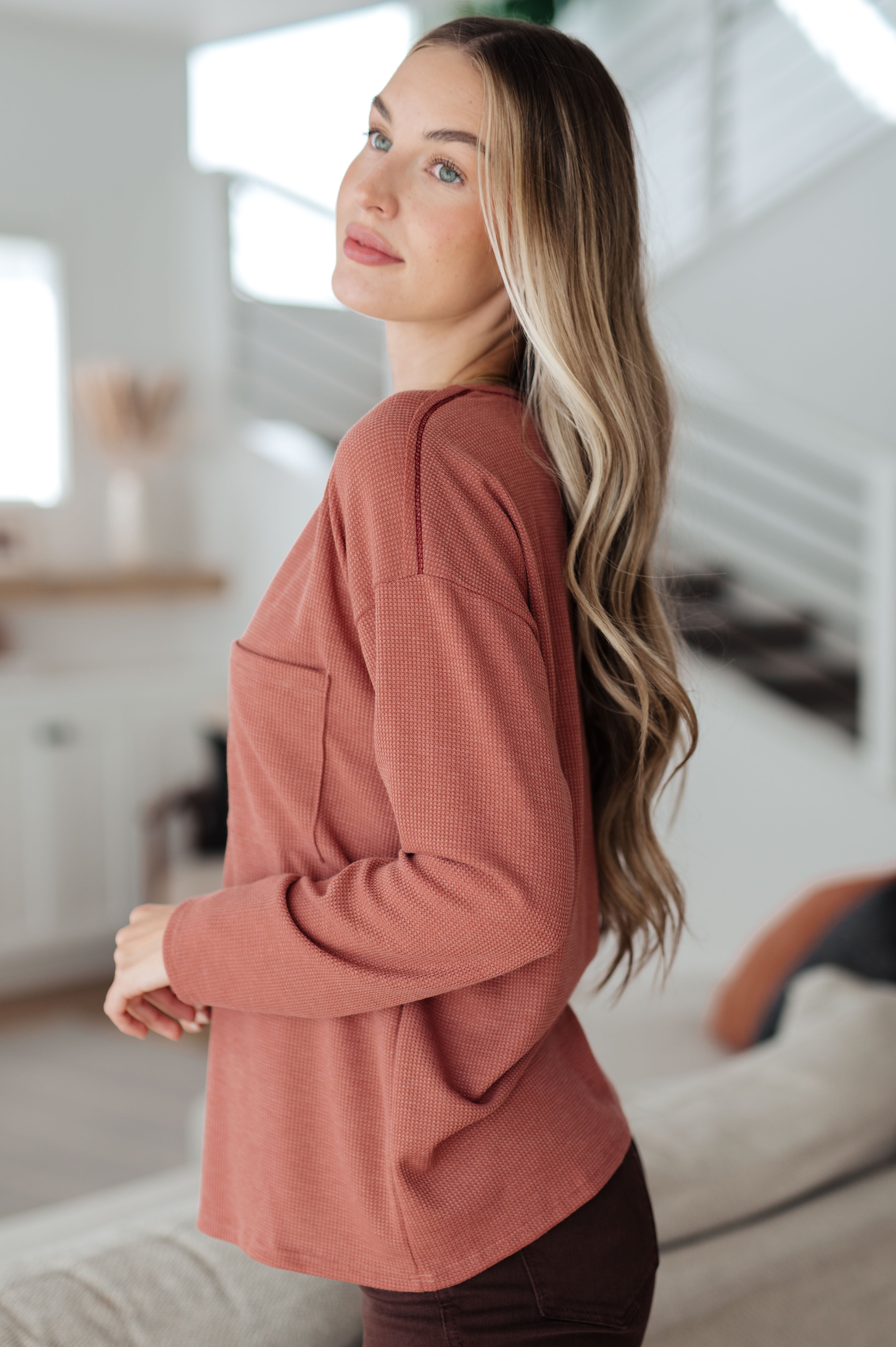 Side view of woman wearing rust-colored Behind The Scenes waffle knit top with pocket in bright modern home setting