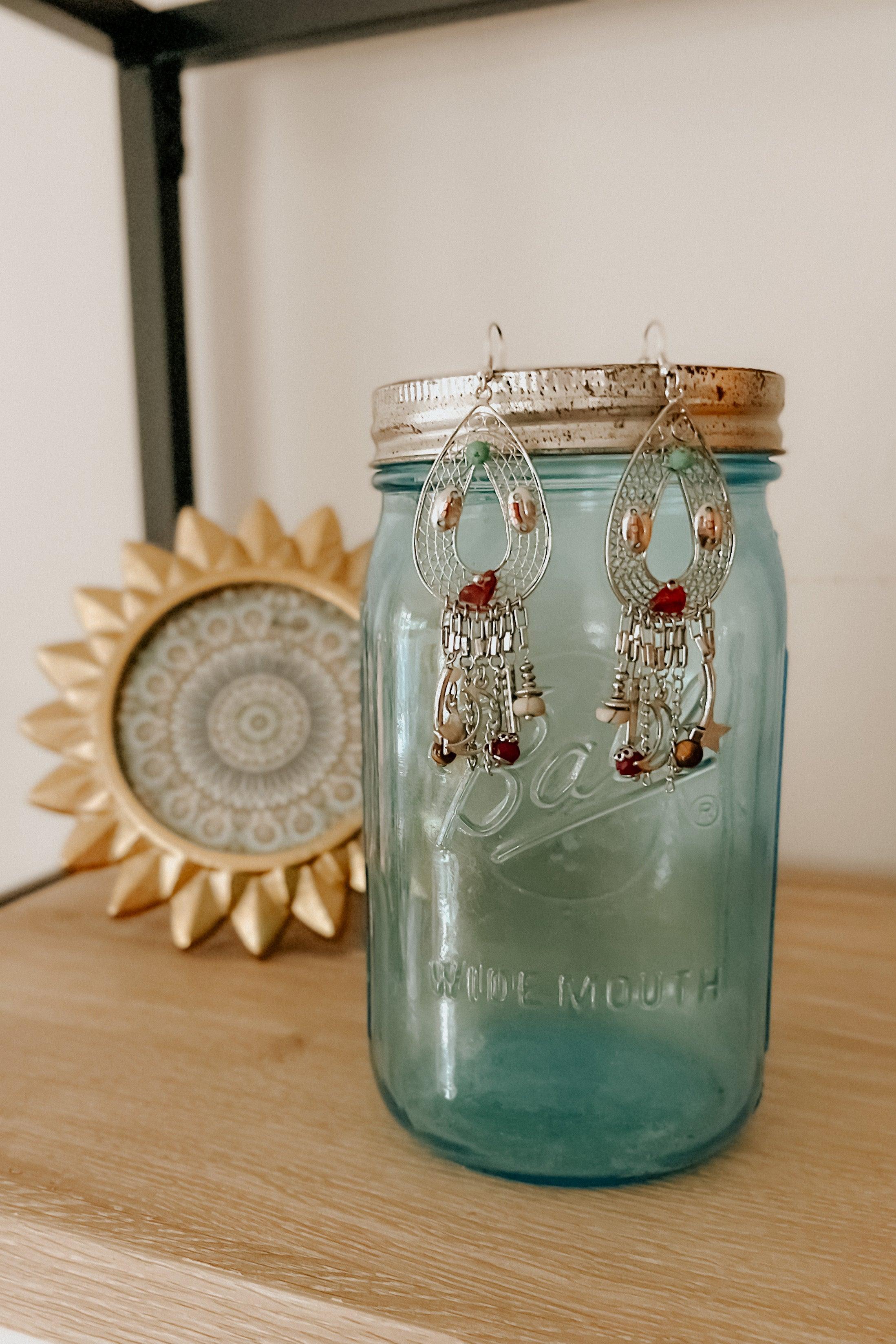 Silver teardrop-shaped earrings with dangling beads in red, brown, and metallic tones displayed on a blue glass jar