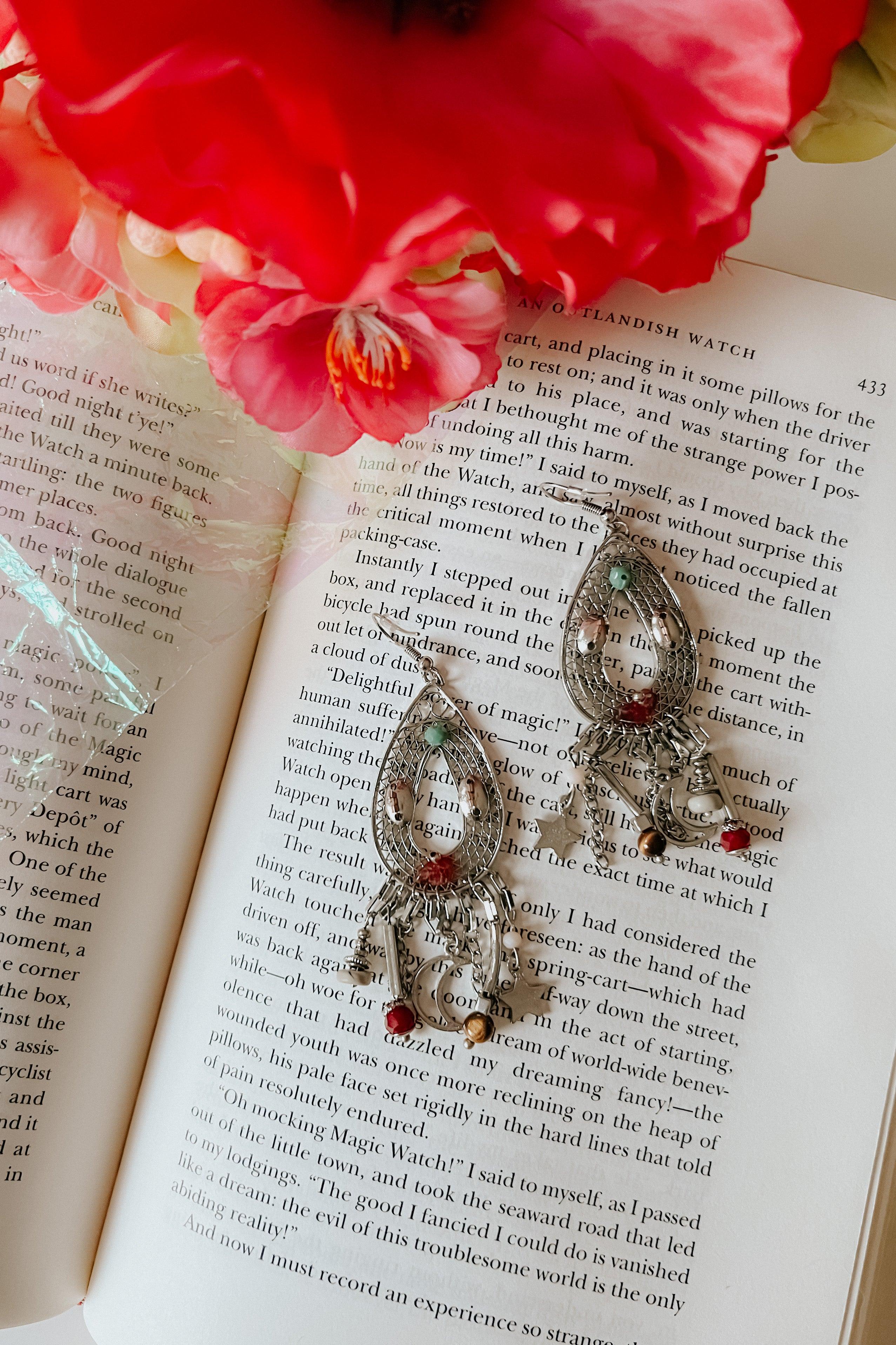 Silver-tone teardrop earrings with charms and colored beads resting on an open book beneath pink and red flowers