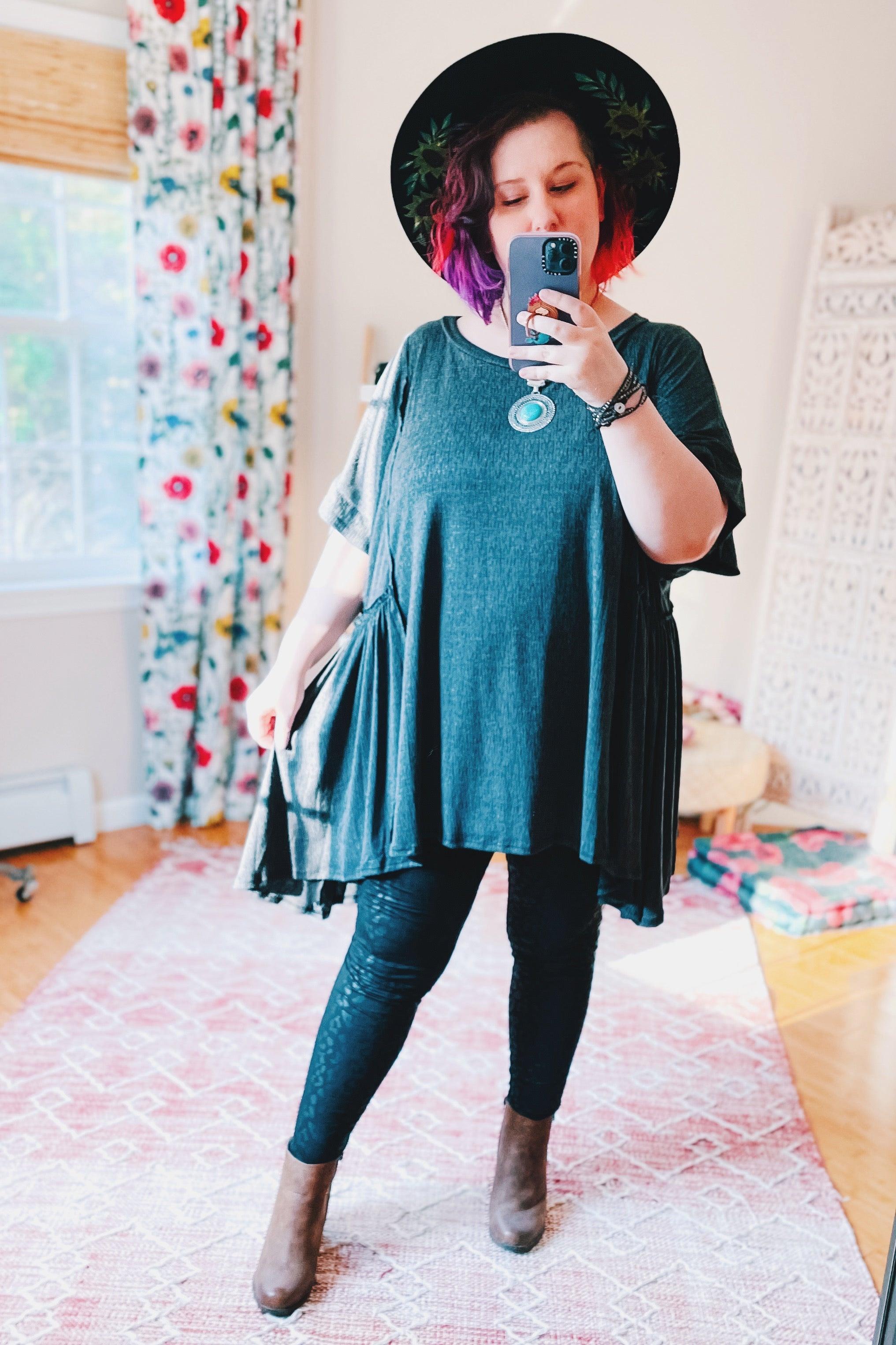Woman taking mirror selfie wearing oversized dark gray shark bite tunic, black hat, patterned leggings, and brown ankle boots indoors