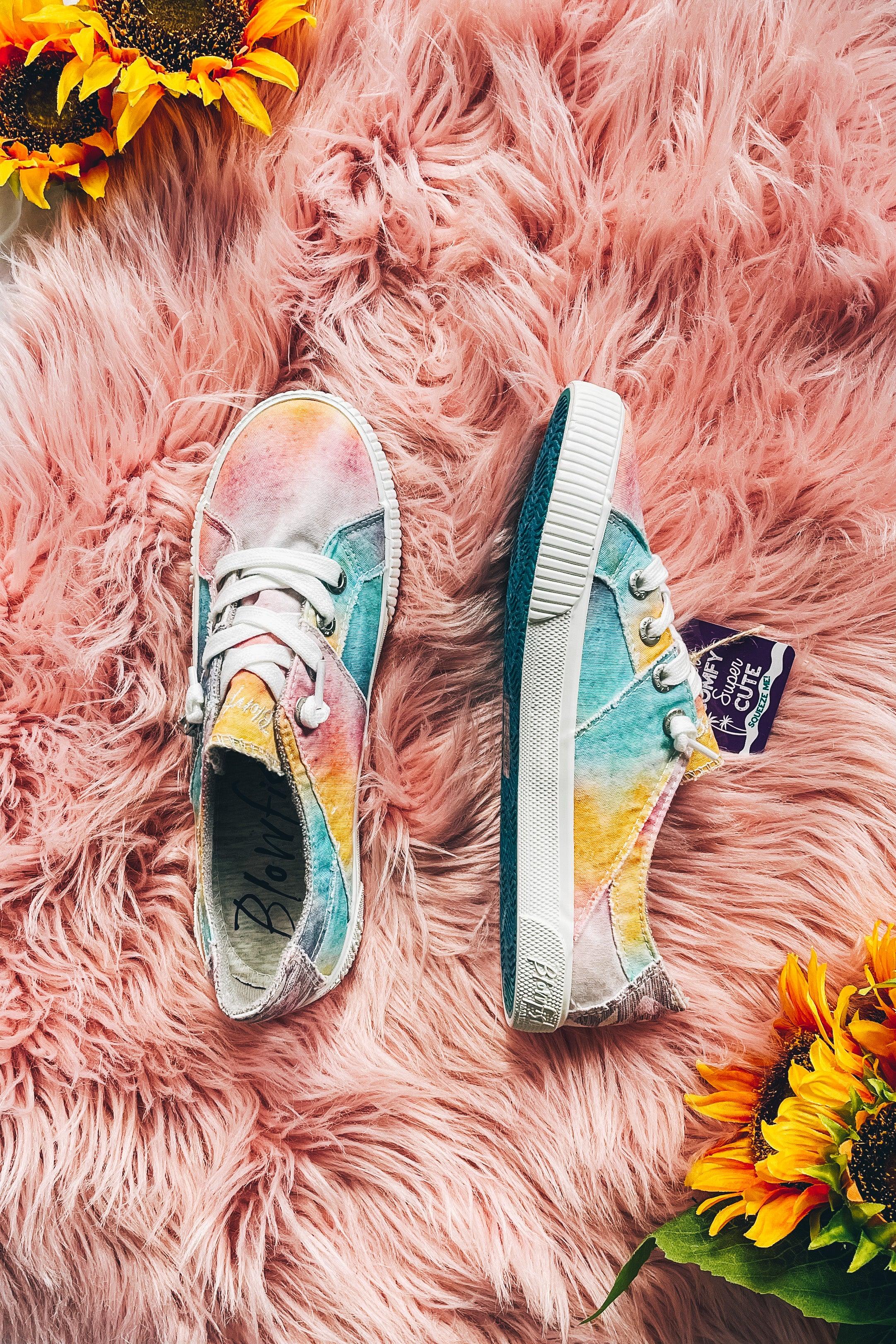 Pair of colorful tie-dye sneakers with white laces on soft pink fluffy rug accented by vibrant sunflowers nearby