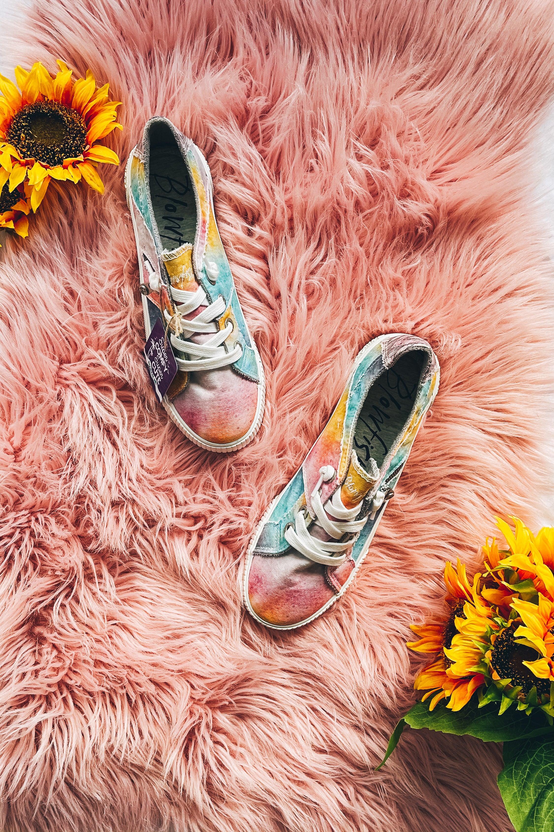 Overhead view of pastel tie-dye Blowfish Malibu sneakers with white laces on fluffy pink rug surrounded by sunflowers