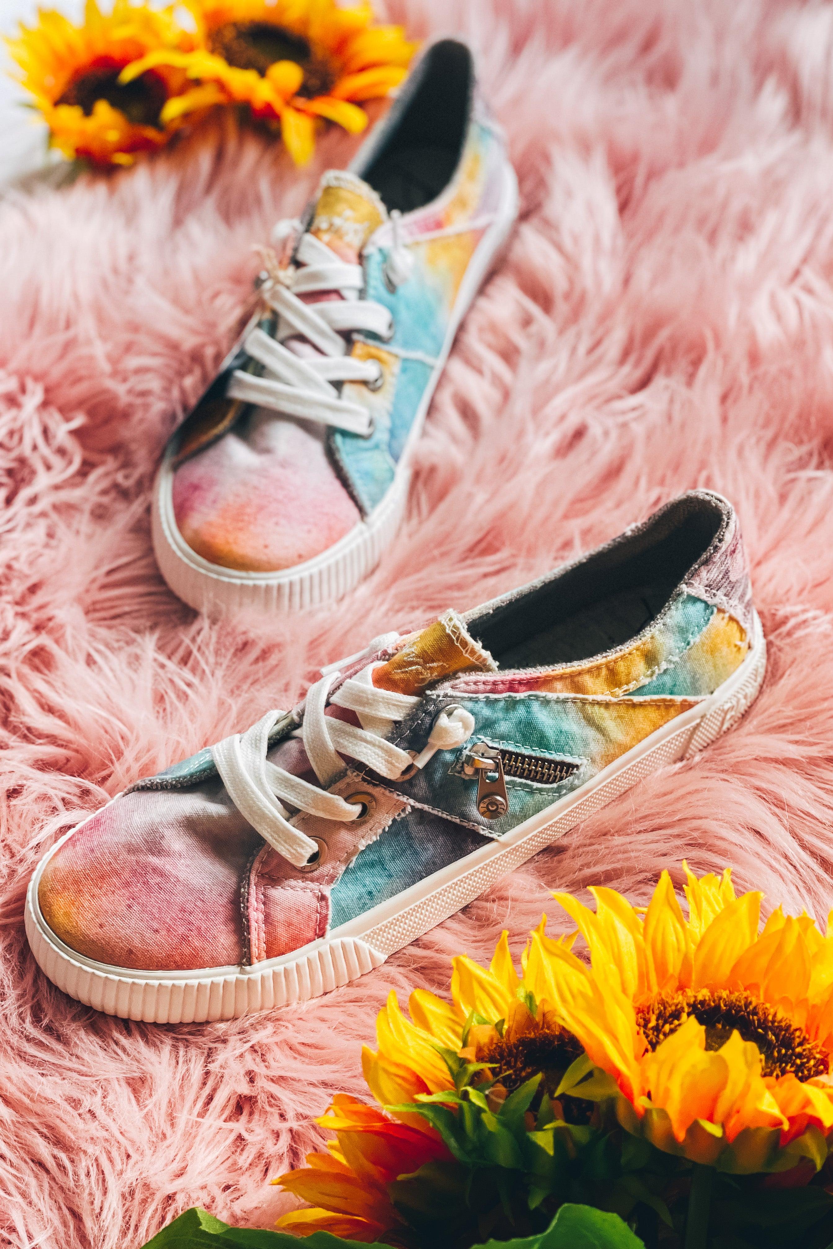 Colorful pastel tie-dye sneakers with white laces and zipper detail on pink fuzzy surface surrounded by sunflowers