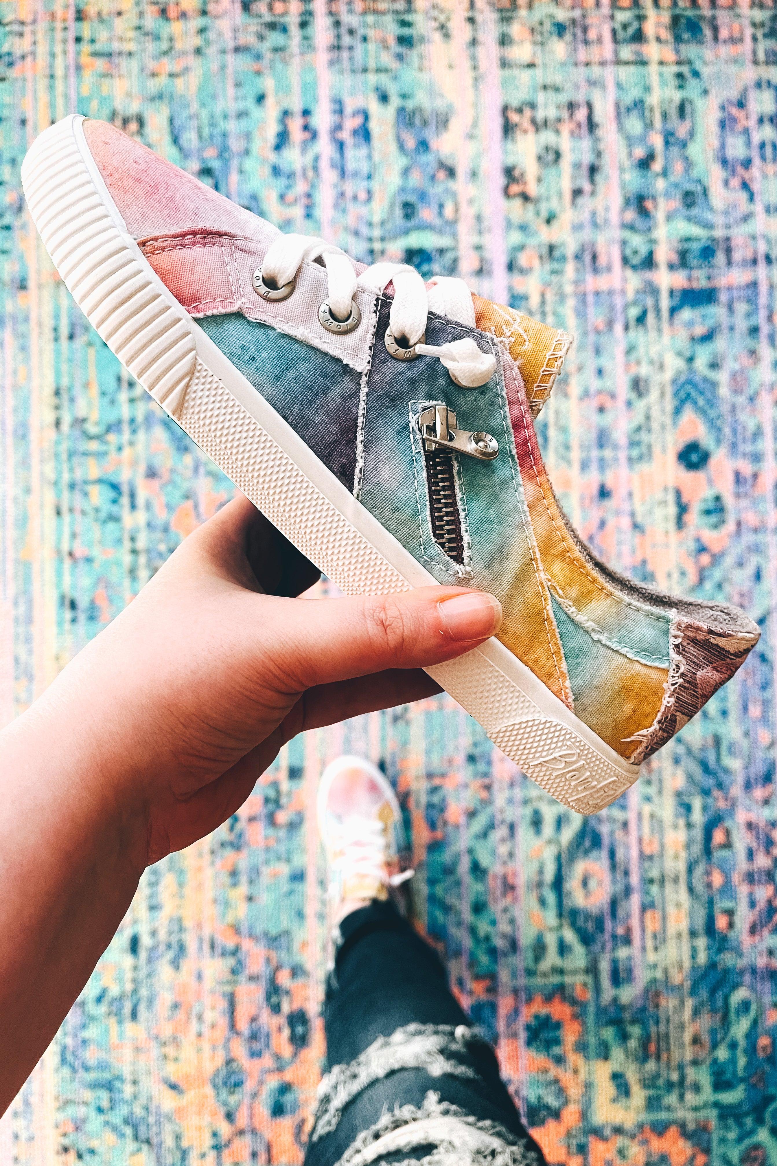 Hand holding multicolor pastel tie-dye sneaker with white laces and decorative zipper over blue patterned rug