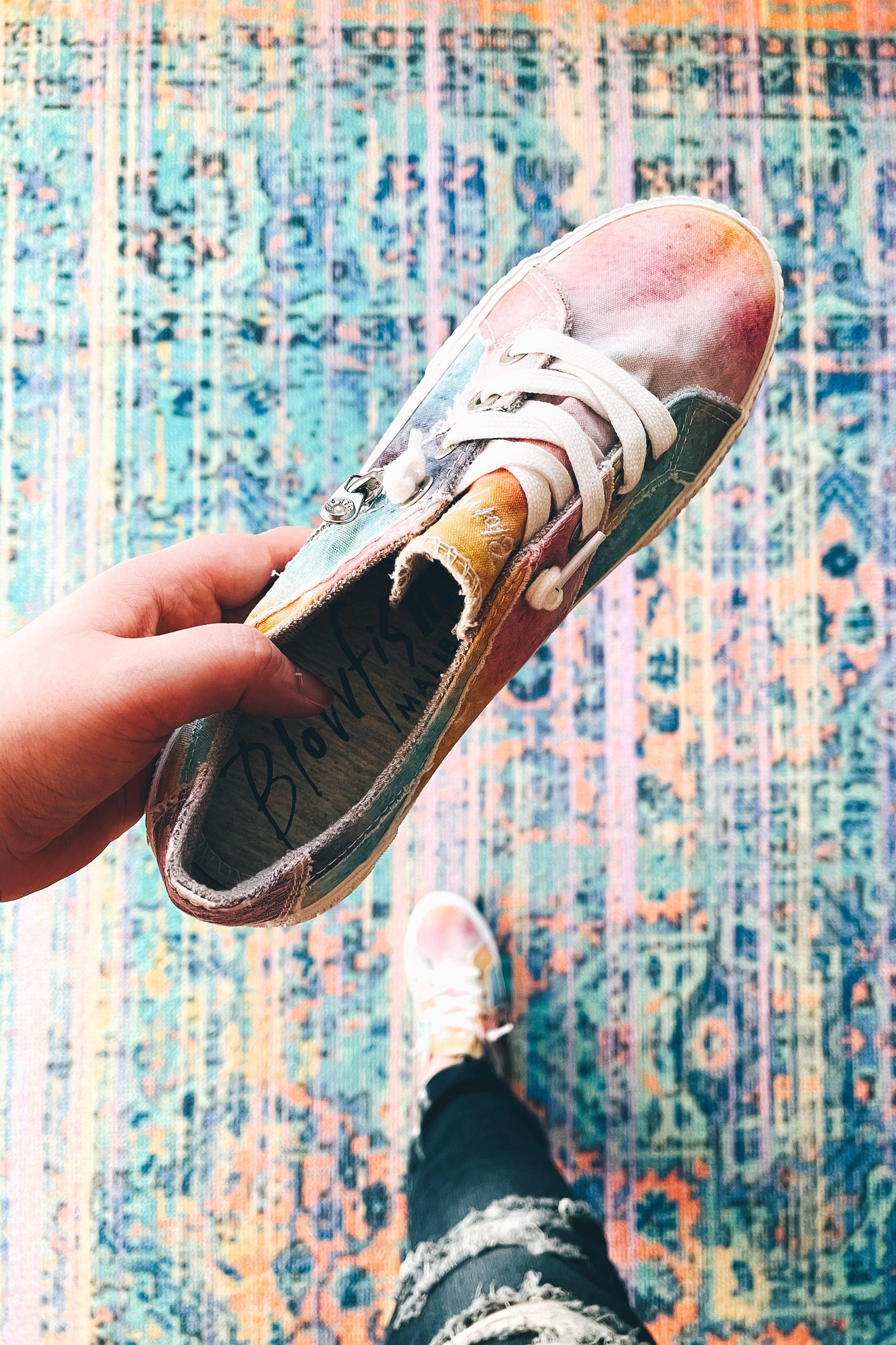 Hand holding a colorful tie-dye sneaker over a vibrant patterned rug with worn denim jeans visible below