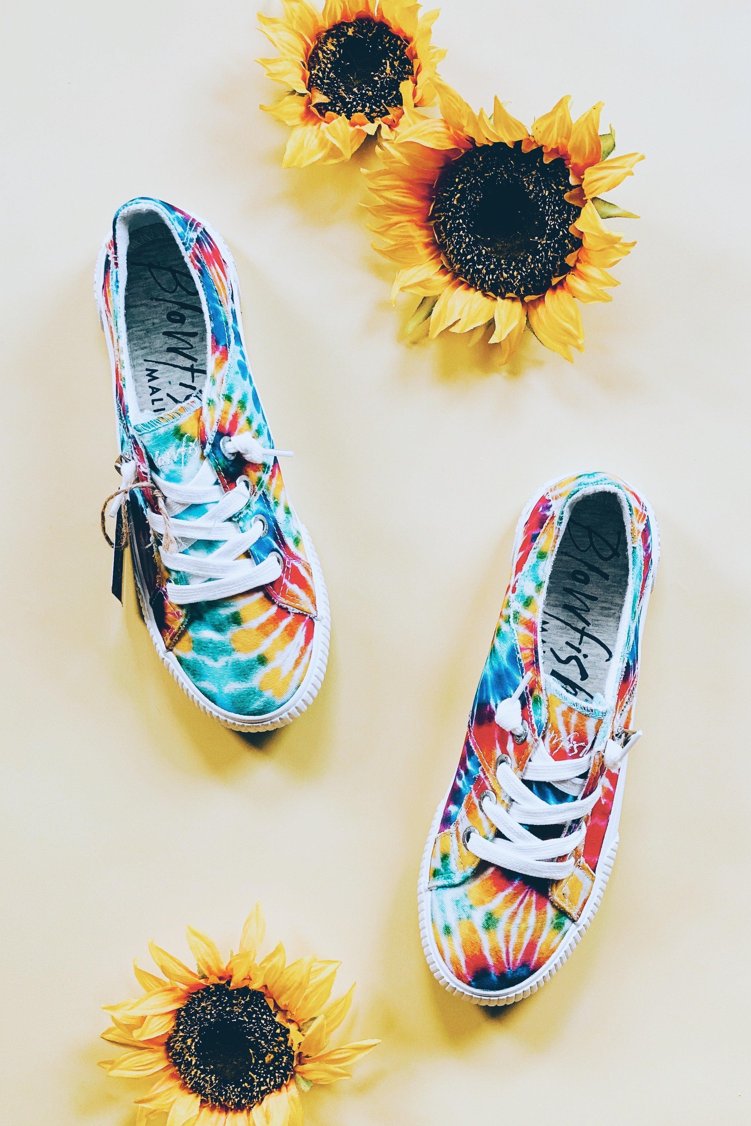 Pair of vibrant rainbow tie-dye canvas sneakers with white laces and rubber soles surrounded by three sunflowers on a light background