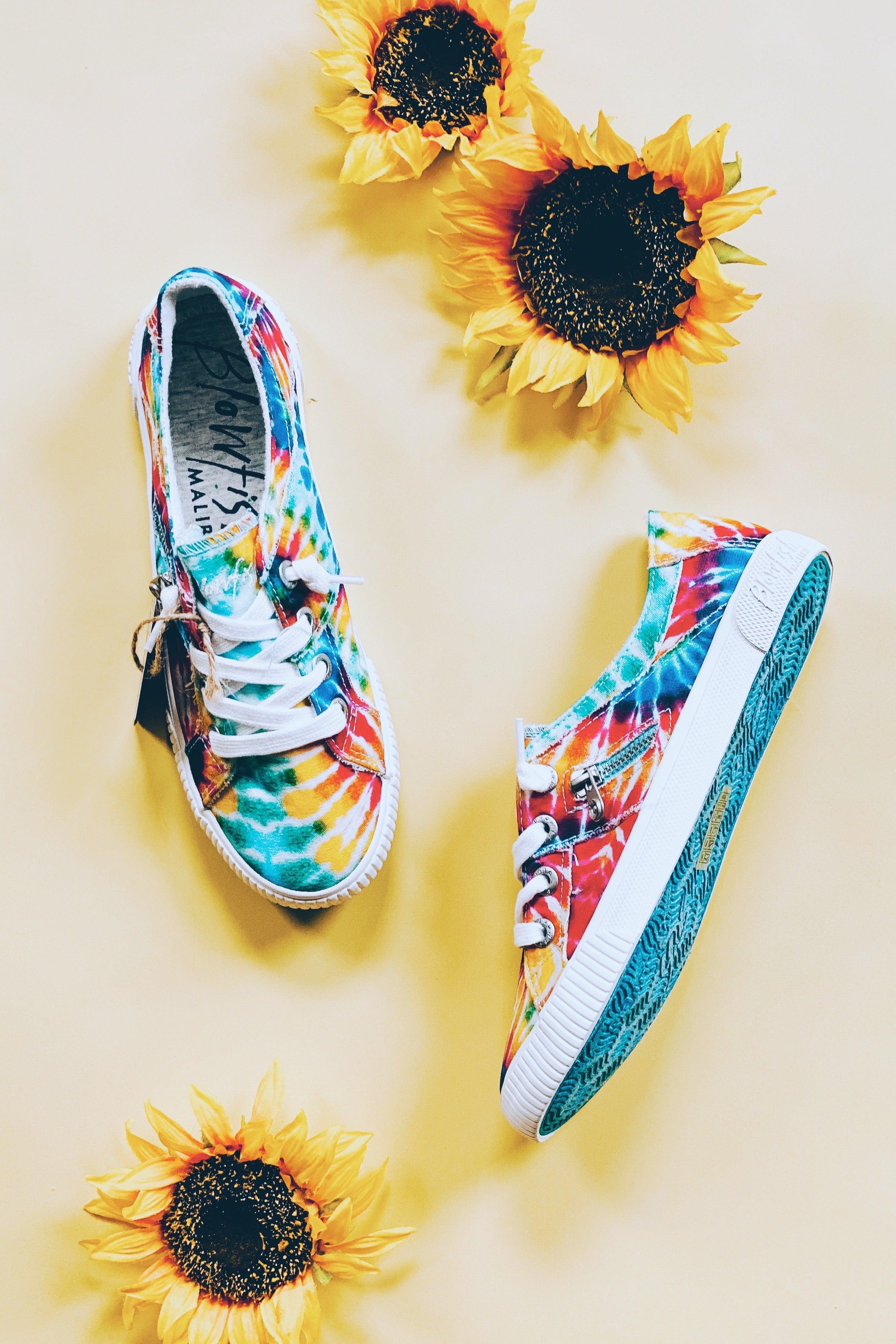 Rainbow tie-dye canvas Blowfish Malibu sneakers with white laces and zipper detail surrounded by three sunflowers on light background