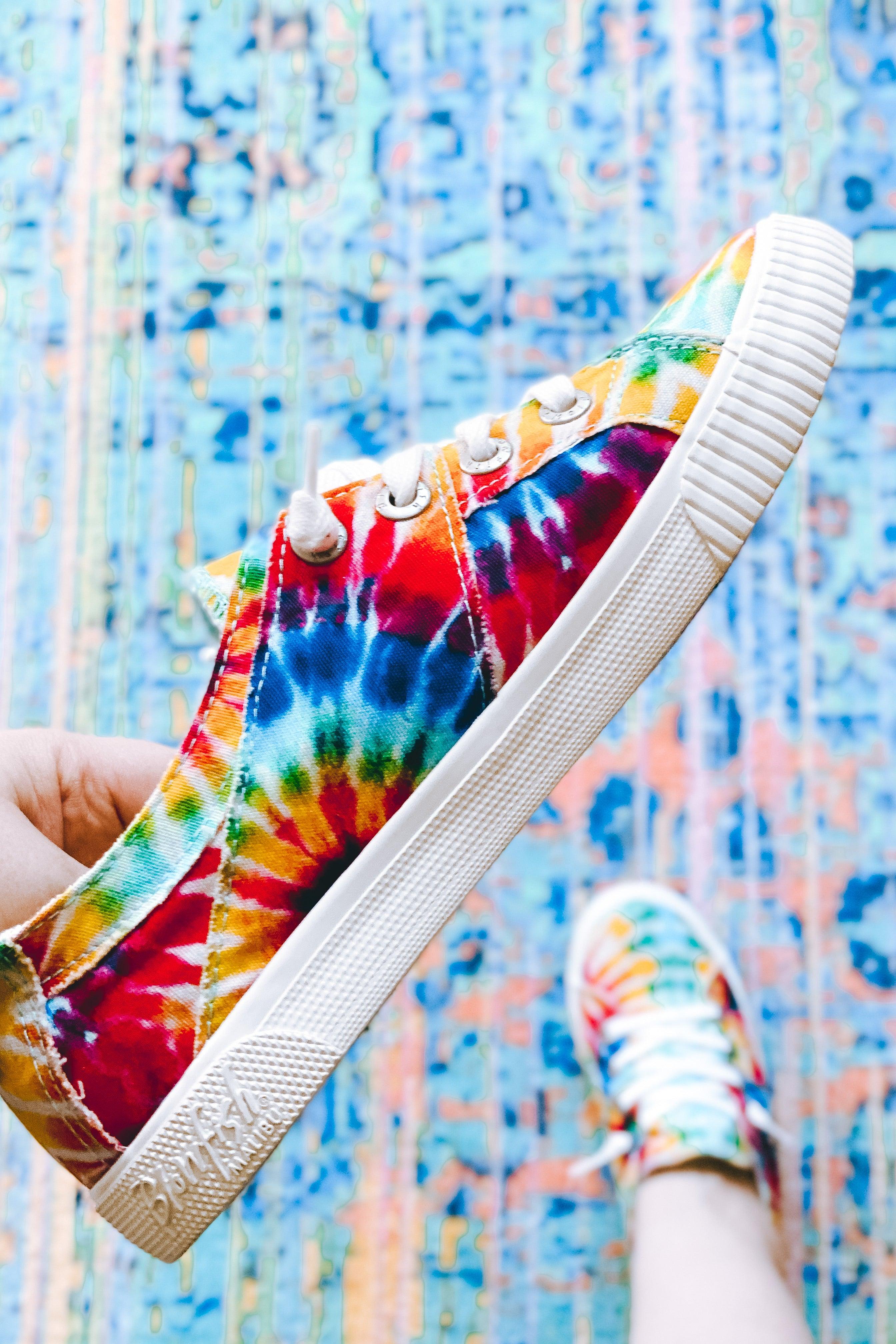 Hand holding Blowfish Malibu rainbow tie-dye canvas sneaker with white laces over colorful patterned floor