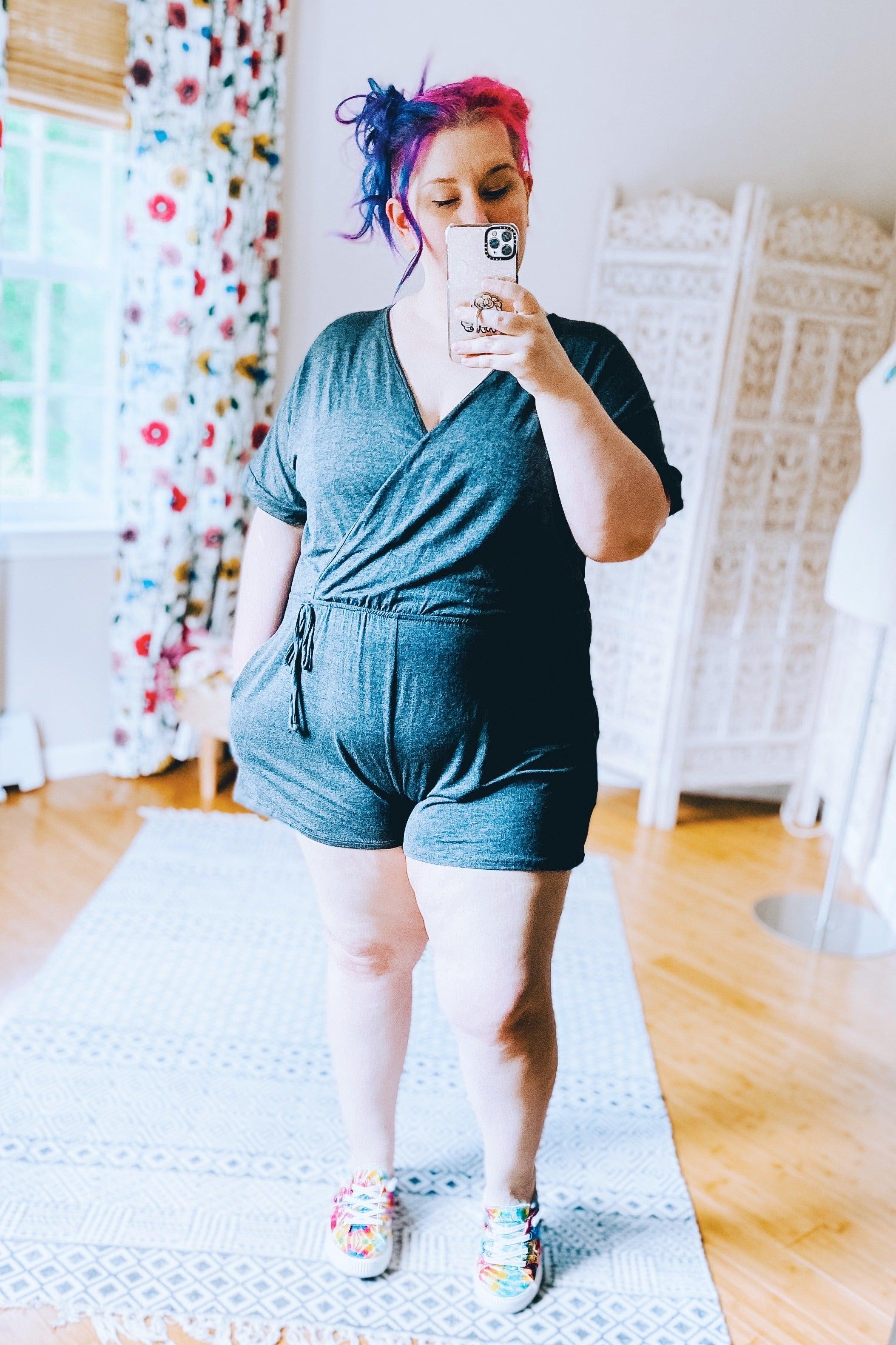Woman with vibrant pink and purple hair wearing a dark gray romper and colorful tie-dye sneakers in bright room