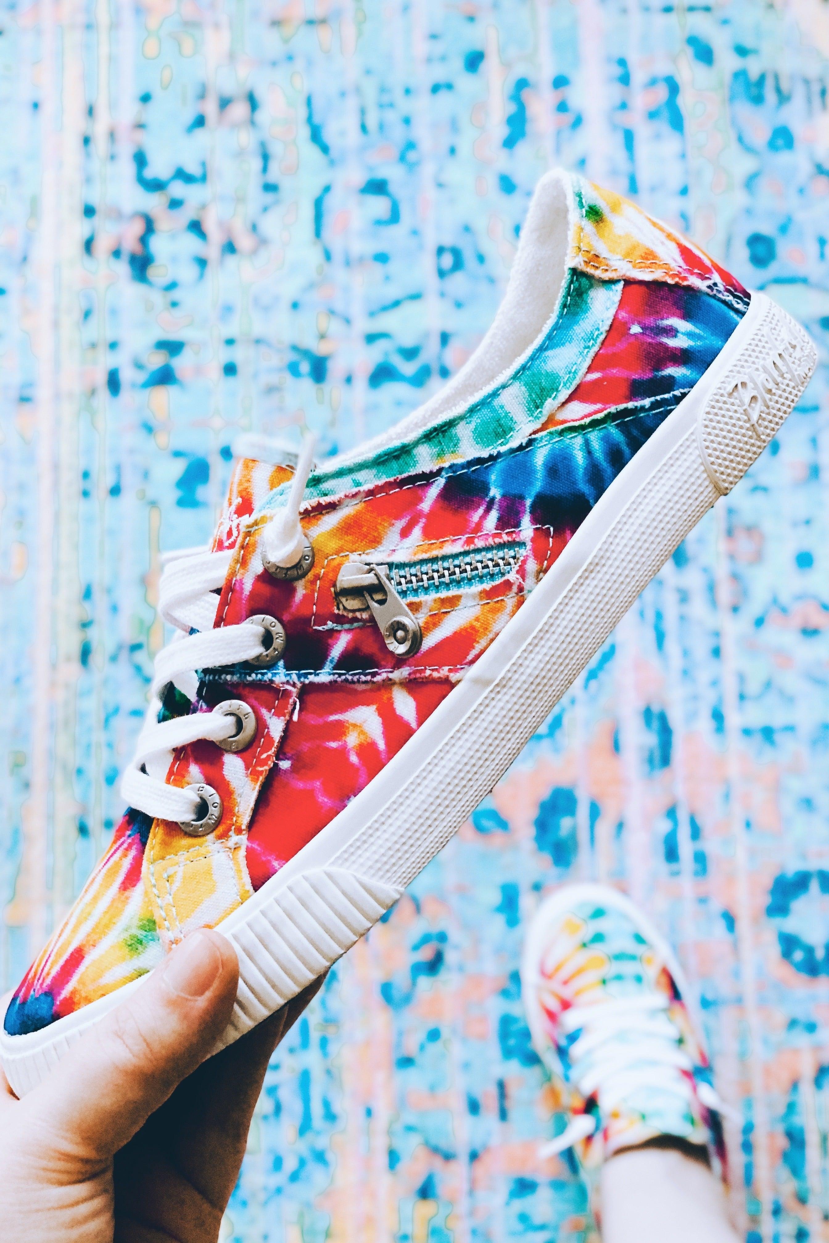 Hand holding vibrant rainbow tie-dye canvas sneaker with white laces and zipper detail over colorful patterned floor