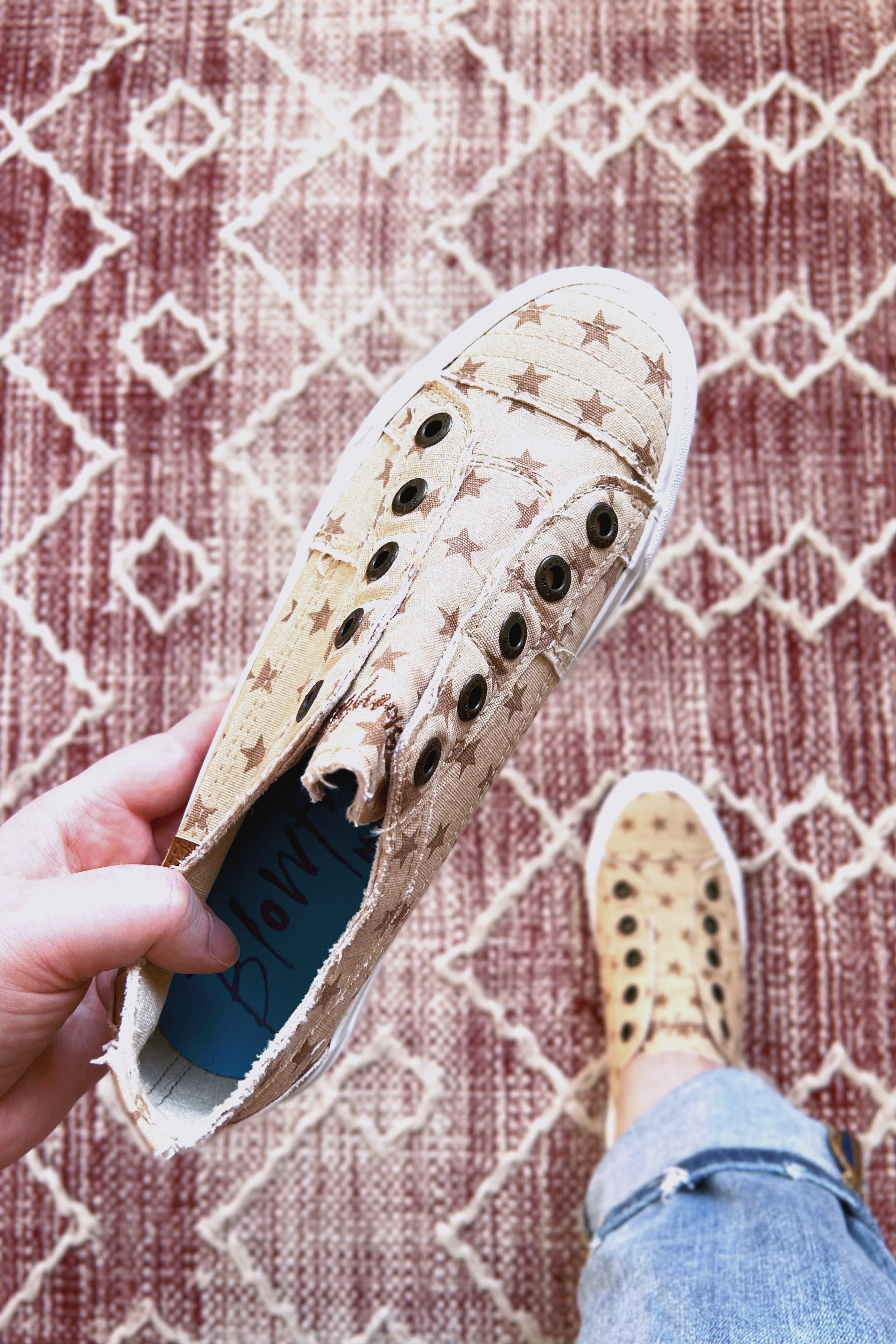 Hand holding beige Blowfish Malibu slip-on sneaker with brown star pattern over red and white geometric rug
