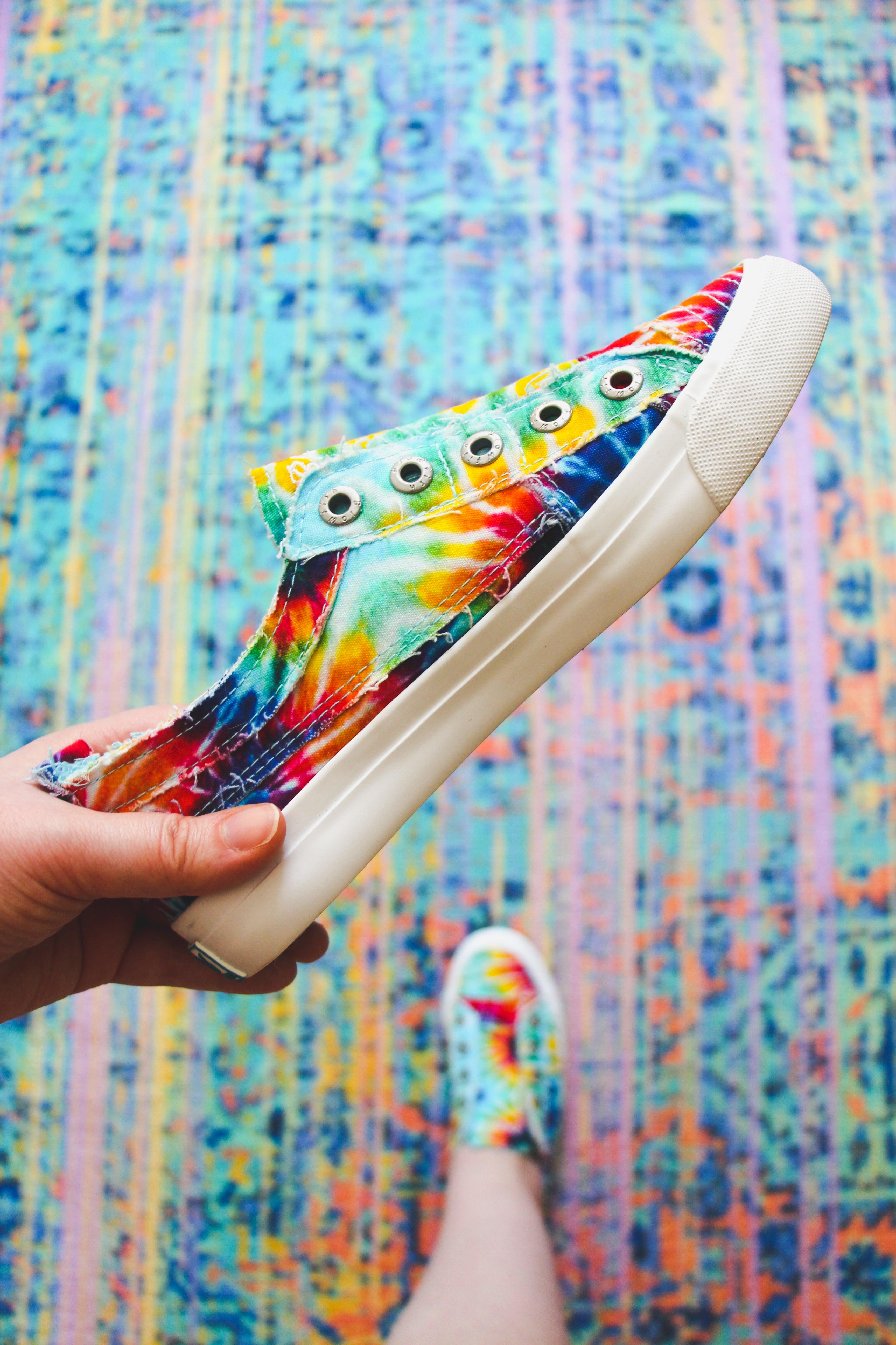 Hand holding rainbow tie-dye canvas sneaker with frayed edges above colorfully patterned rug and matching shoe worn on foot