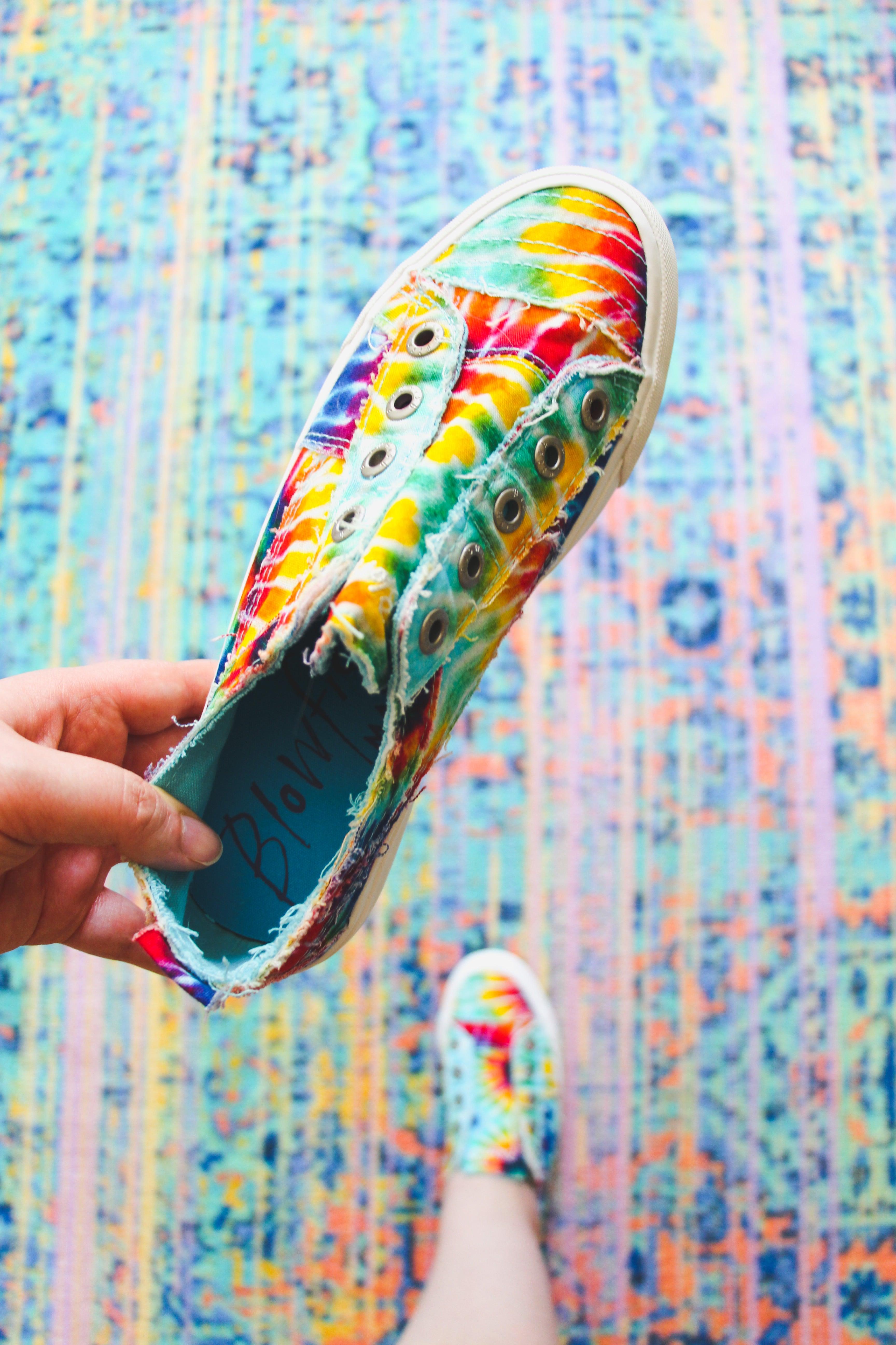 Hand holding rainbow tie-dye canvas sneaker with frayed edges above matching shoe worn on patterned rug