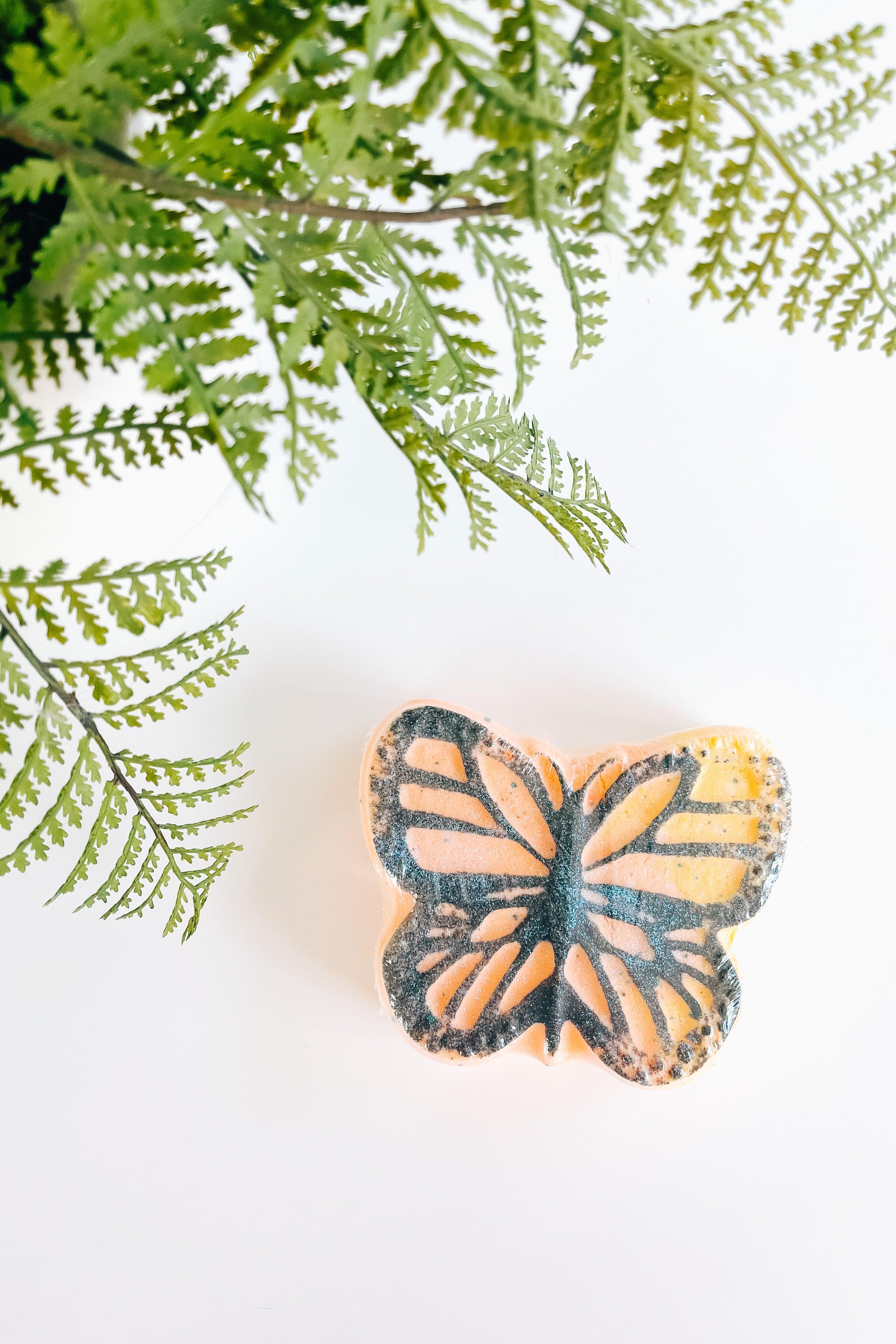 Butterfly-shaped bath bomb with orange, yellow, and black colors next to green fern leaves on white background