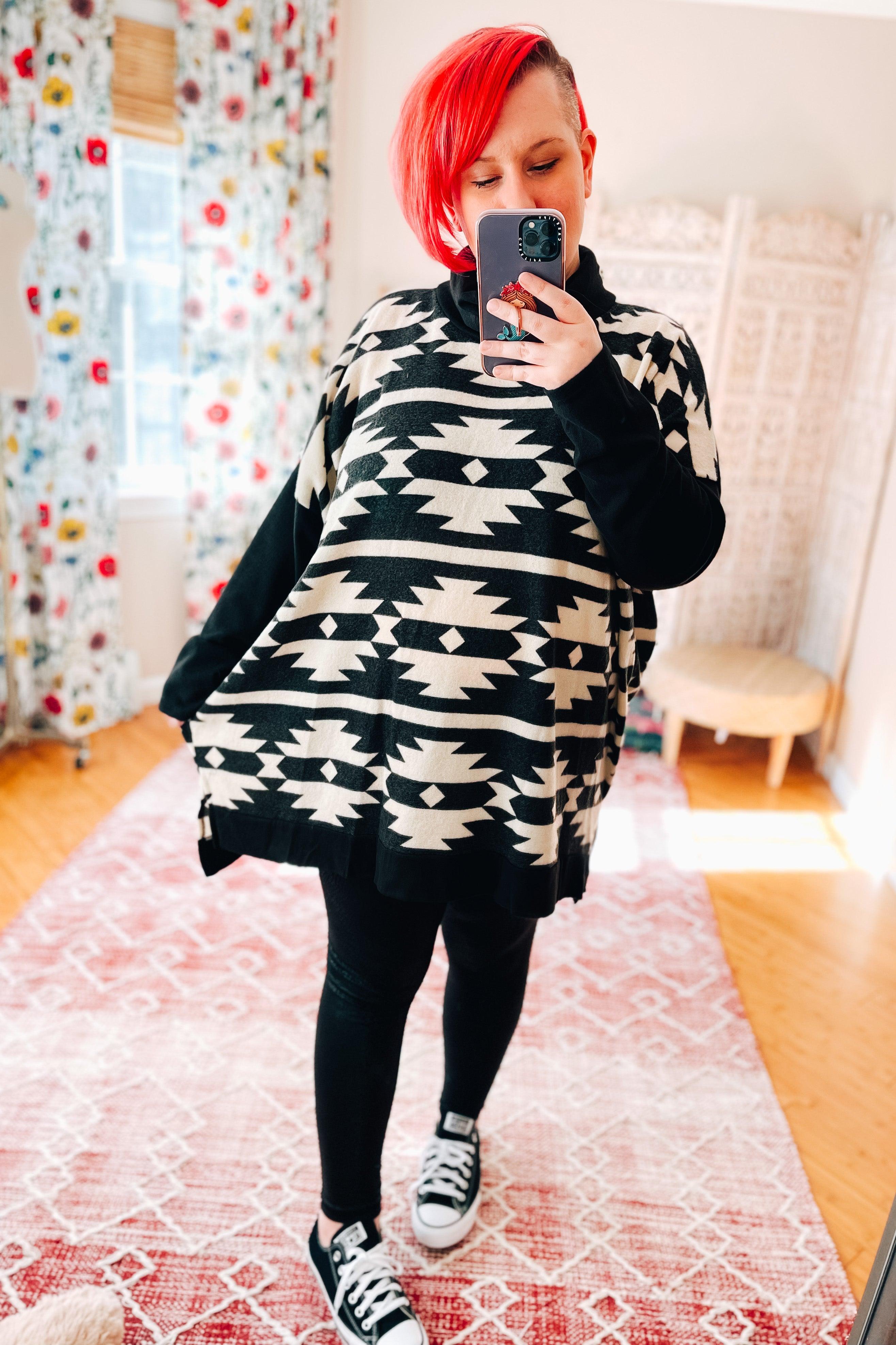Woman with red hair wearing black and white geometric patterned cowl neck pullover with black leggings and sneakers indoors taking selfie