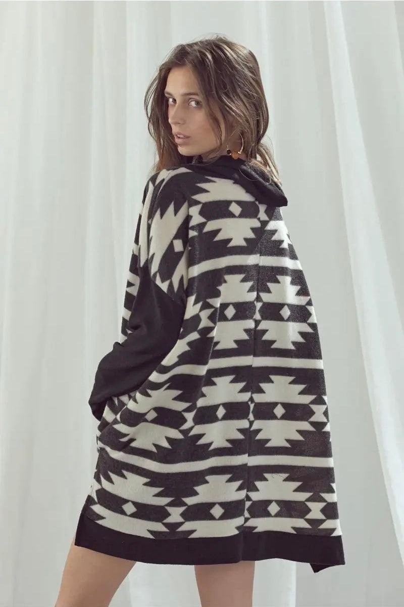 Woman wearing oversized black and white geometric patterned pullover with hood standing sideways against white curtain background