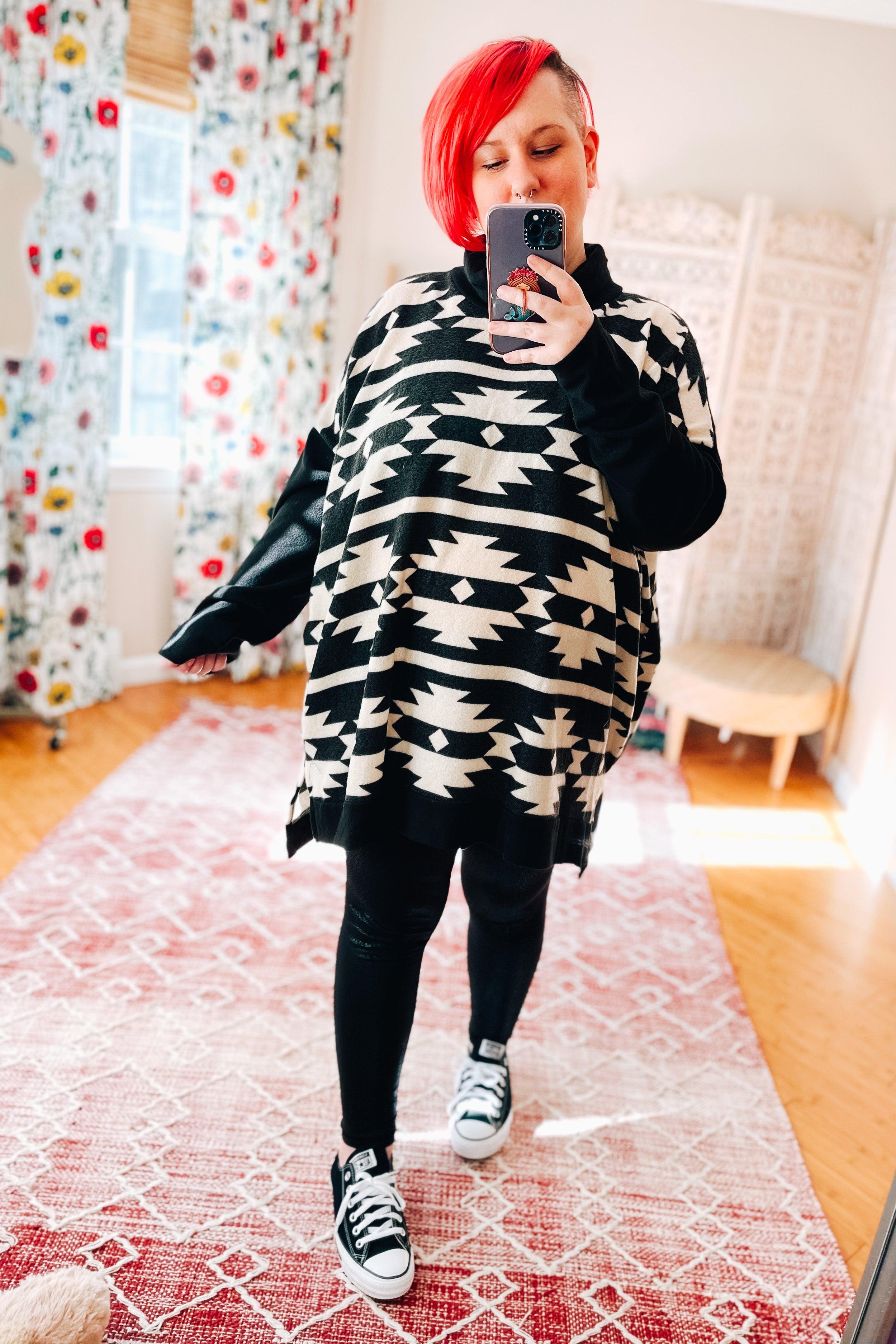 Woman wearing black and white geometric patterned cowl neck pullover with black leggings and sneakers indoors taking selfie