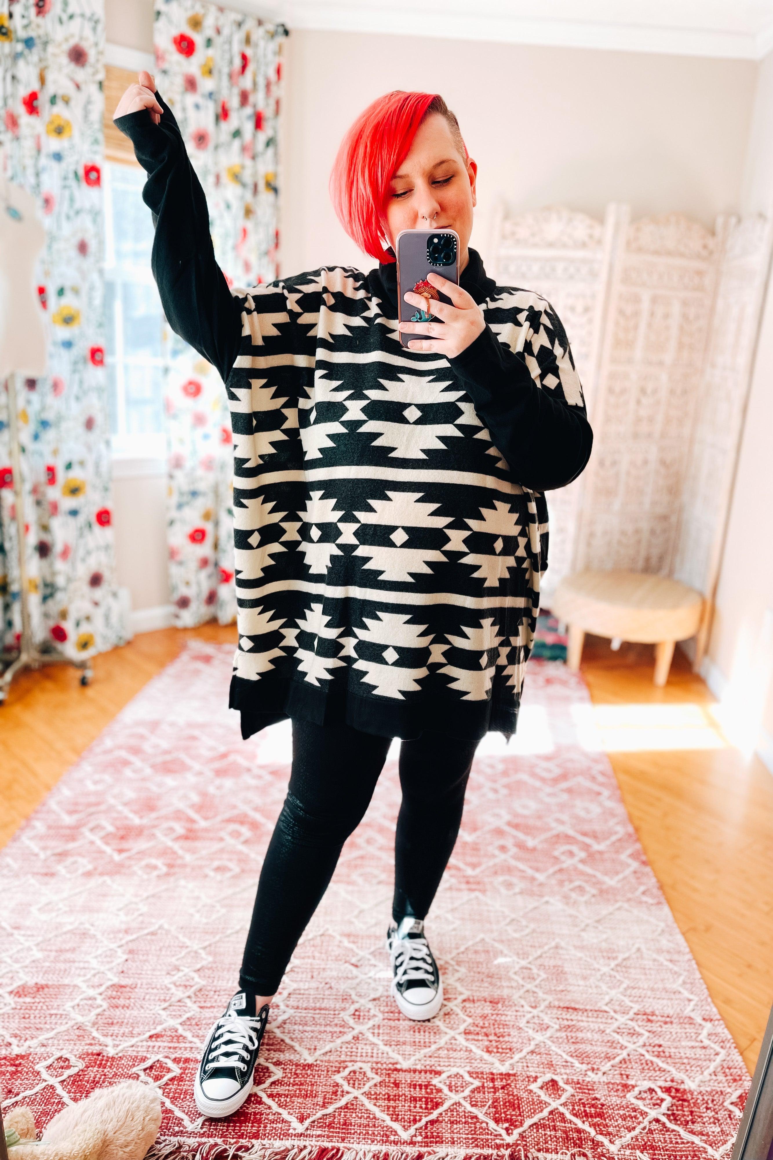 Woman with red hair wearing oversized black and white geometric patterned pullover with black leggings and sneakers taking selfie indoors